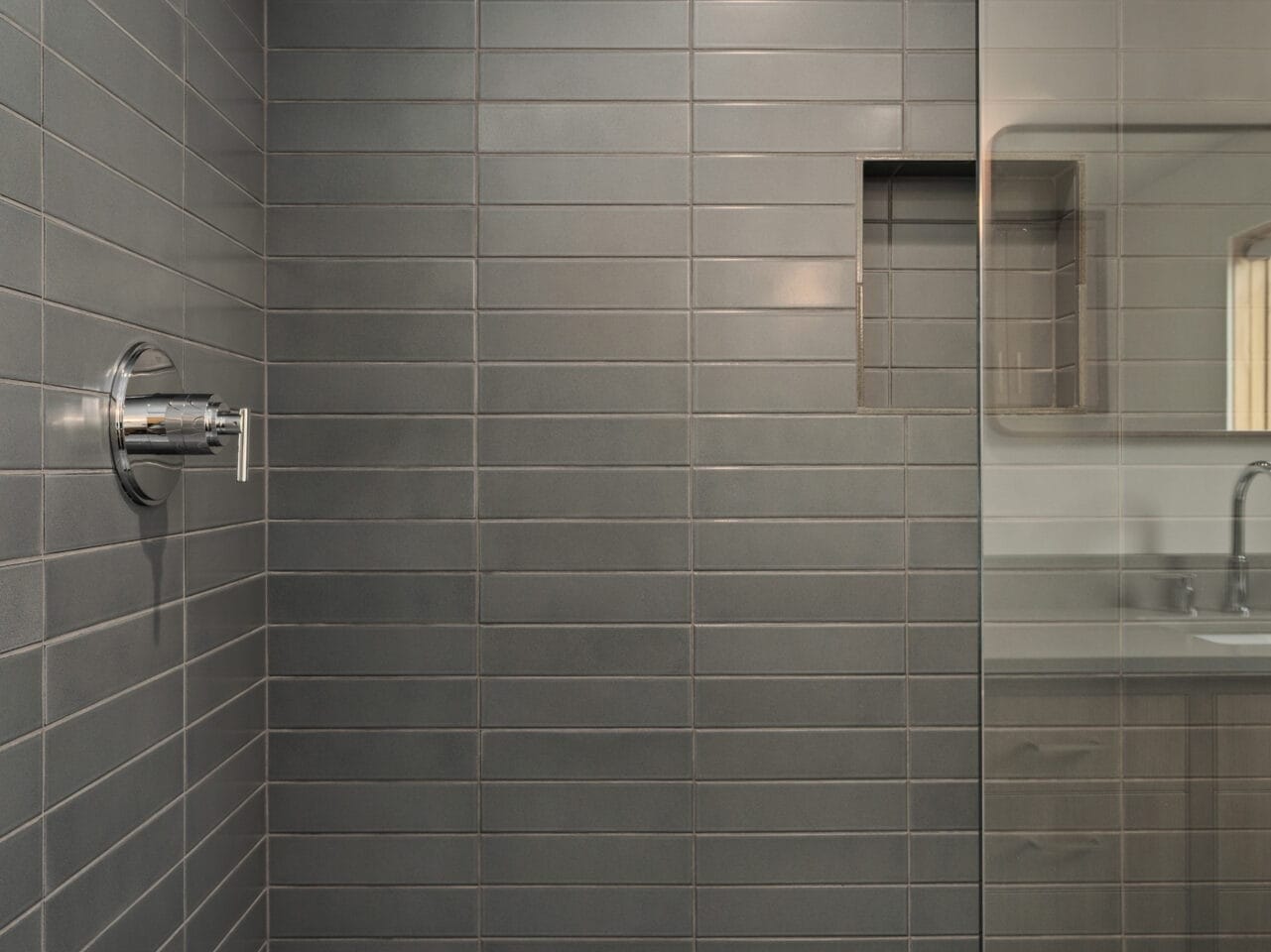 A modern shower in Portland, Oregon, featuring dark gray rectangular wall tiles and a built-in niche. The showerhead is mounted on the left wall, with a glass partition separating it from the rest of the bathroom. The floor boasts an elegant herringbone pattern.