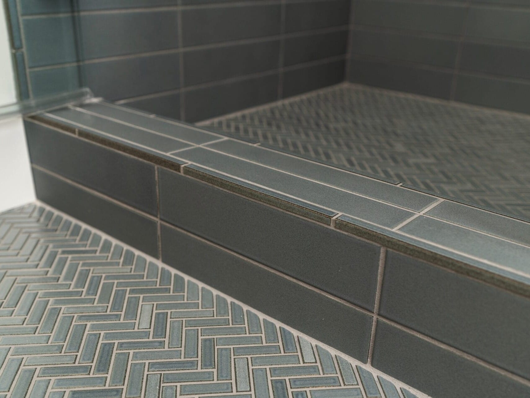 Close-up of a shower floor in a Portland, Oregon home, showcasing elegant dark green tiles arranged in a herringbone pattern. Matching rectangular tiles on the edge and wall complete this modern and stylish design.