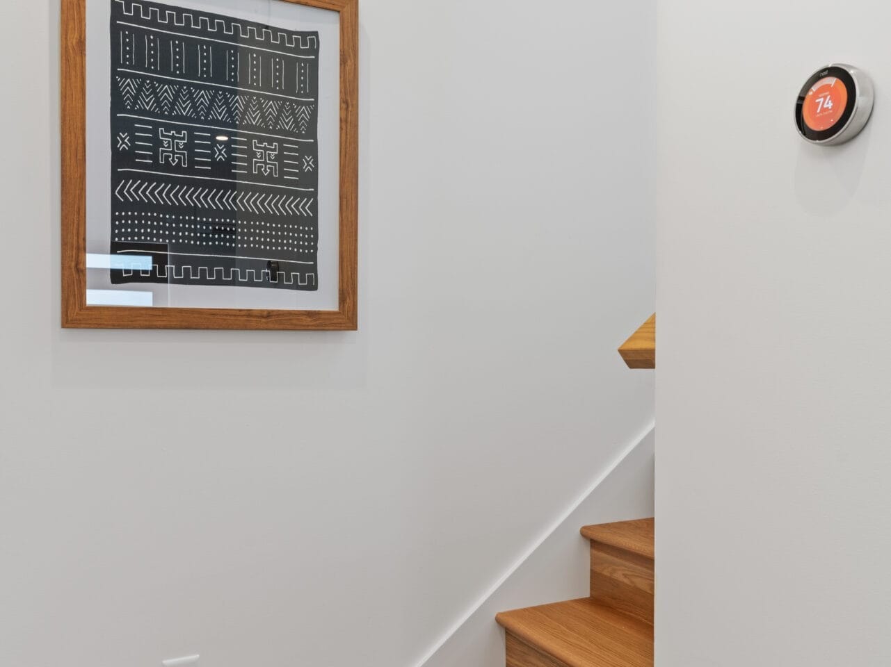 A wooden staircase ascends against a white wall in the heart of Portland, Oregon. A framed black and white geometric art piece graces the wall, while a round digital thermostat displays 74 degrees nearby.