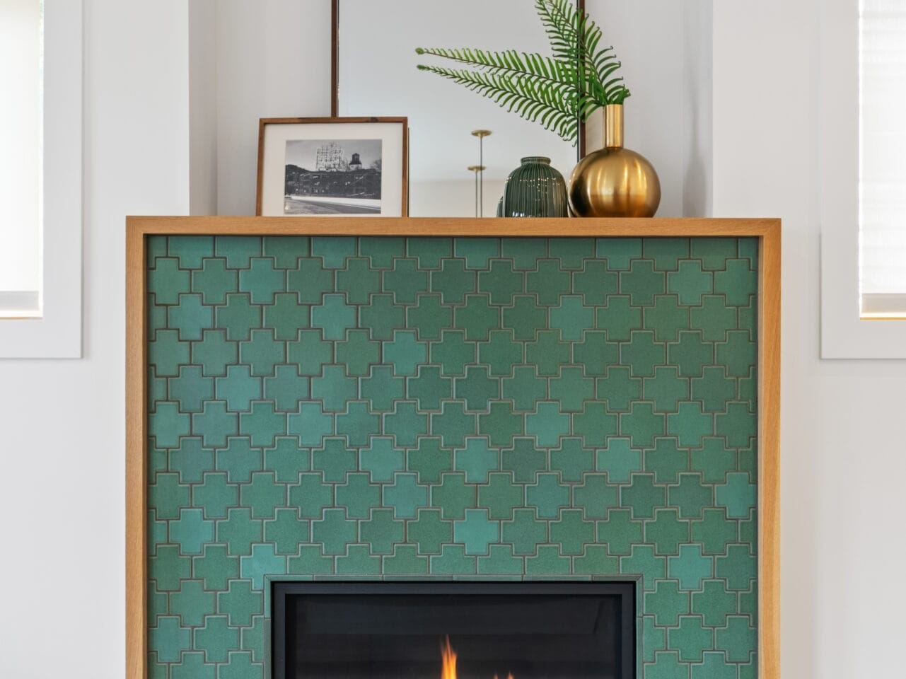 A cozy living room in a Portland, Oregon home features a green tiled fireplace with a flickering fire. A wooden mantel holds a mirror, green vase, gold spherical vase, and framed photo. In front, a round wooden coffee table displays seashells and books.