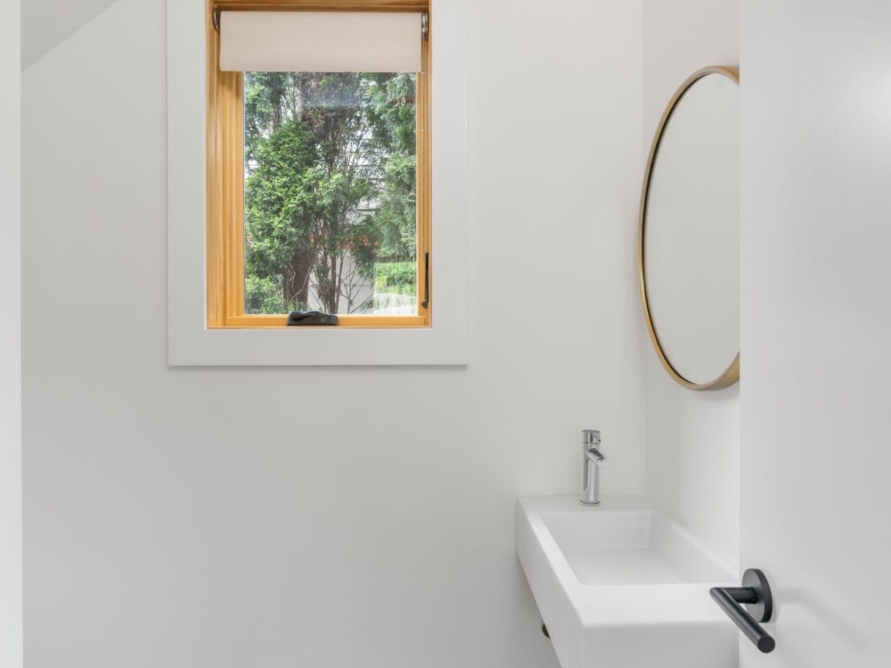 A minimalist bathroom in Portland, Oregon, features a wall-mounted sink with a round mirror above it. A single light bulb hangs from a cord next to the mirror. A small window with a wooden frame reveals lush greenery outside. The floor is polished concrete, and the walls are white.