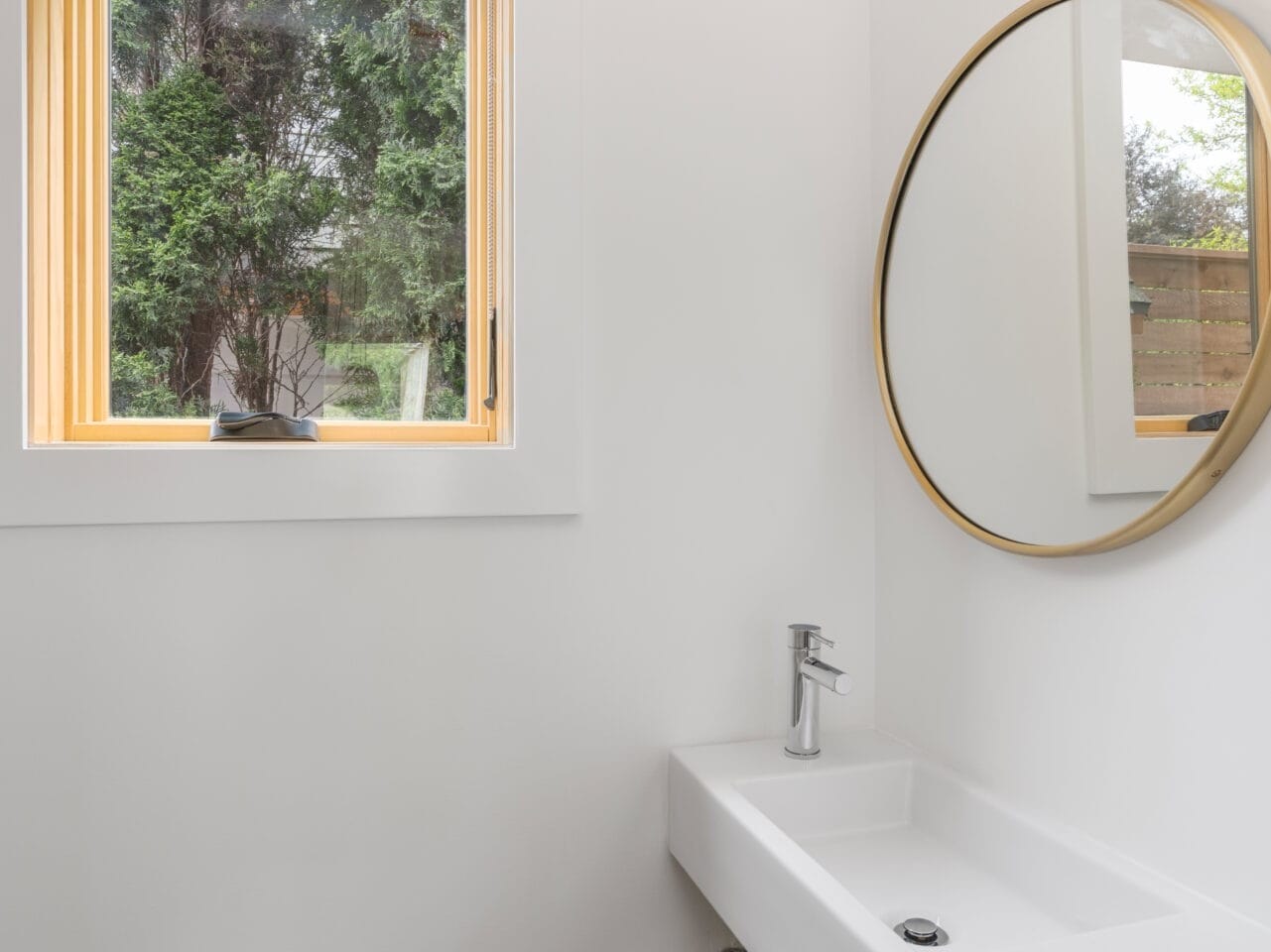 A minimalist Portland, Oregon bathroom features a white rectangular sink and round mirror. Natural light floods in through a wooden-framed window revealing greenery outside, complemented by a small pendant light hanging gracefully above.