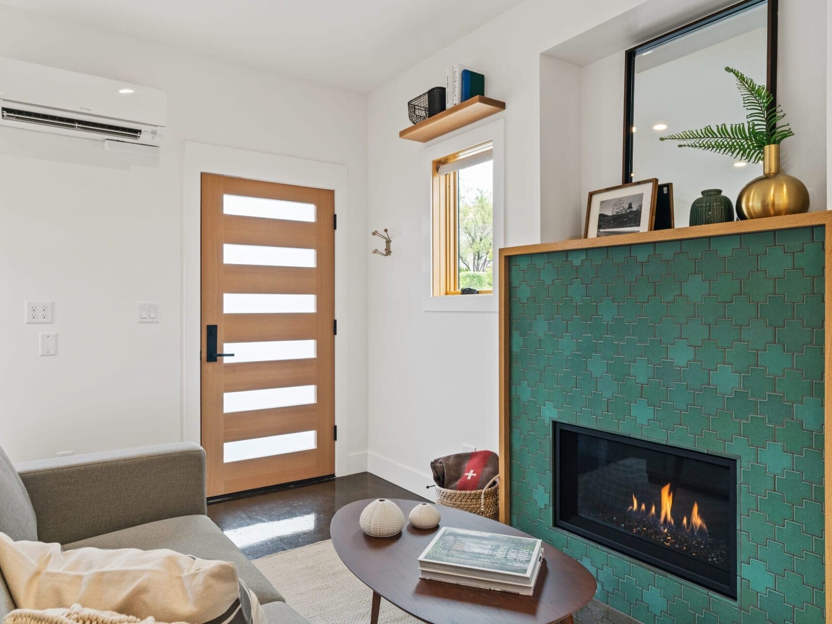 A cozy living room in Portland, Oregon, features a gray sofa, a teal-tiled fireplace, and a wooden door with horizontal glass panels. A small window above the fireplace showcases potted plants and framed photos. A woven basket and decor items adorn a round coffee table.