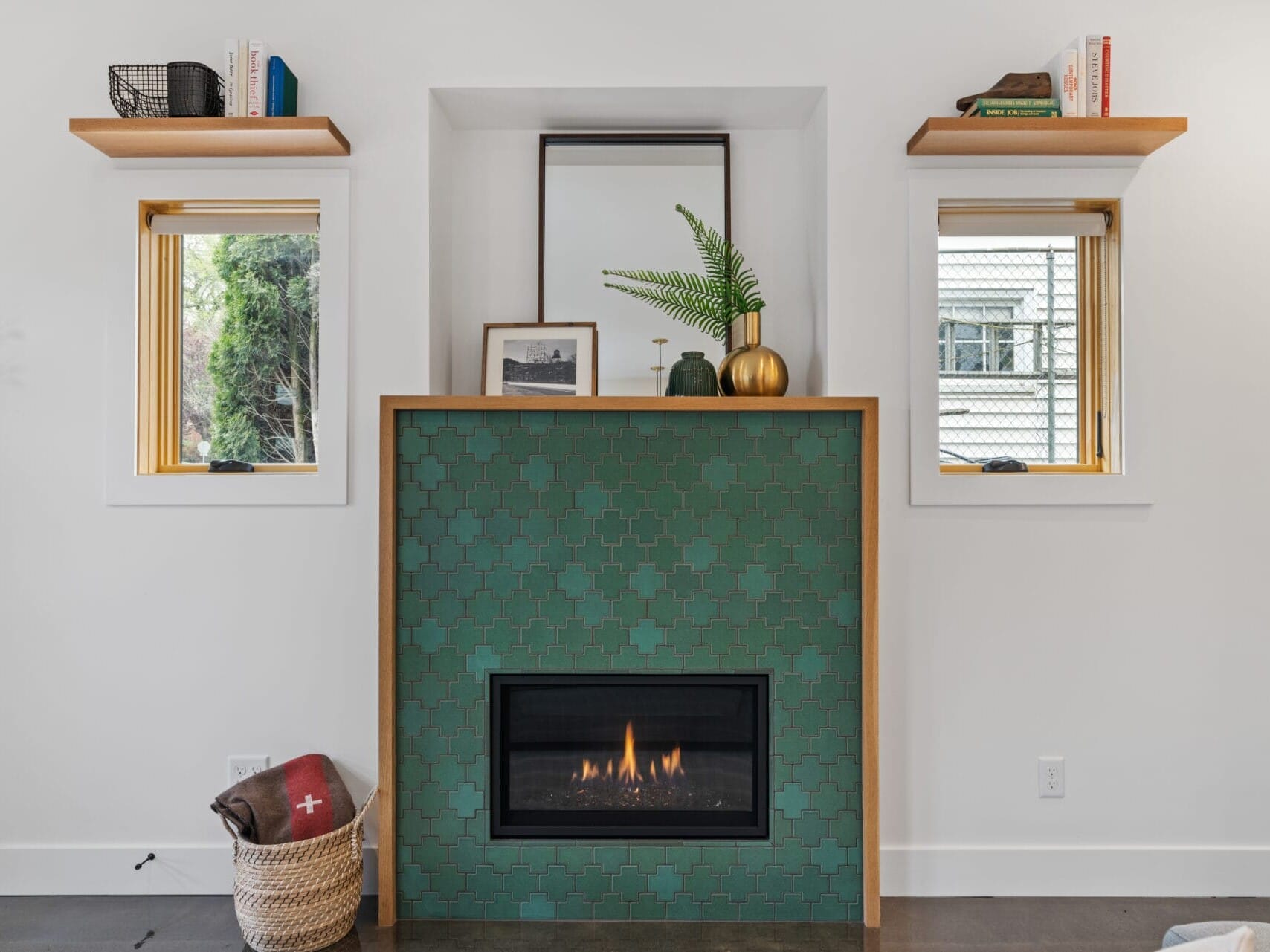 A cozy living room in Portland, Oregon, features a green tiled fireplace adorned with decorative items on the mantel. Two small windows flank the fireplace, each with wooden shelves. A basket with a blanket is placed on the floor nearby.