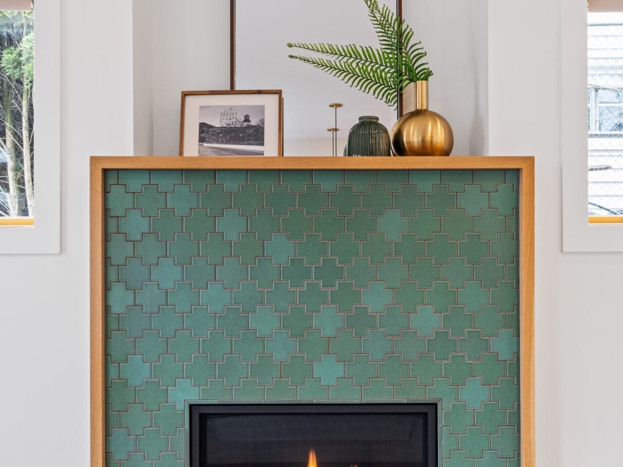 A modern fireplace with a green tiled pattern and wooden mantel captures the essence of Portland, Oregon. On top, there's a framed photo, a square mirror, a green vase, and a gold vase with fern leaves. Flanking it are windows with light wooden shelves that embrace the city's natural vibe.