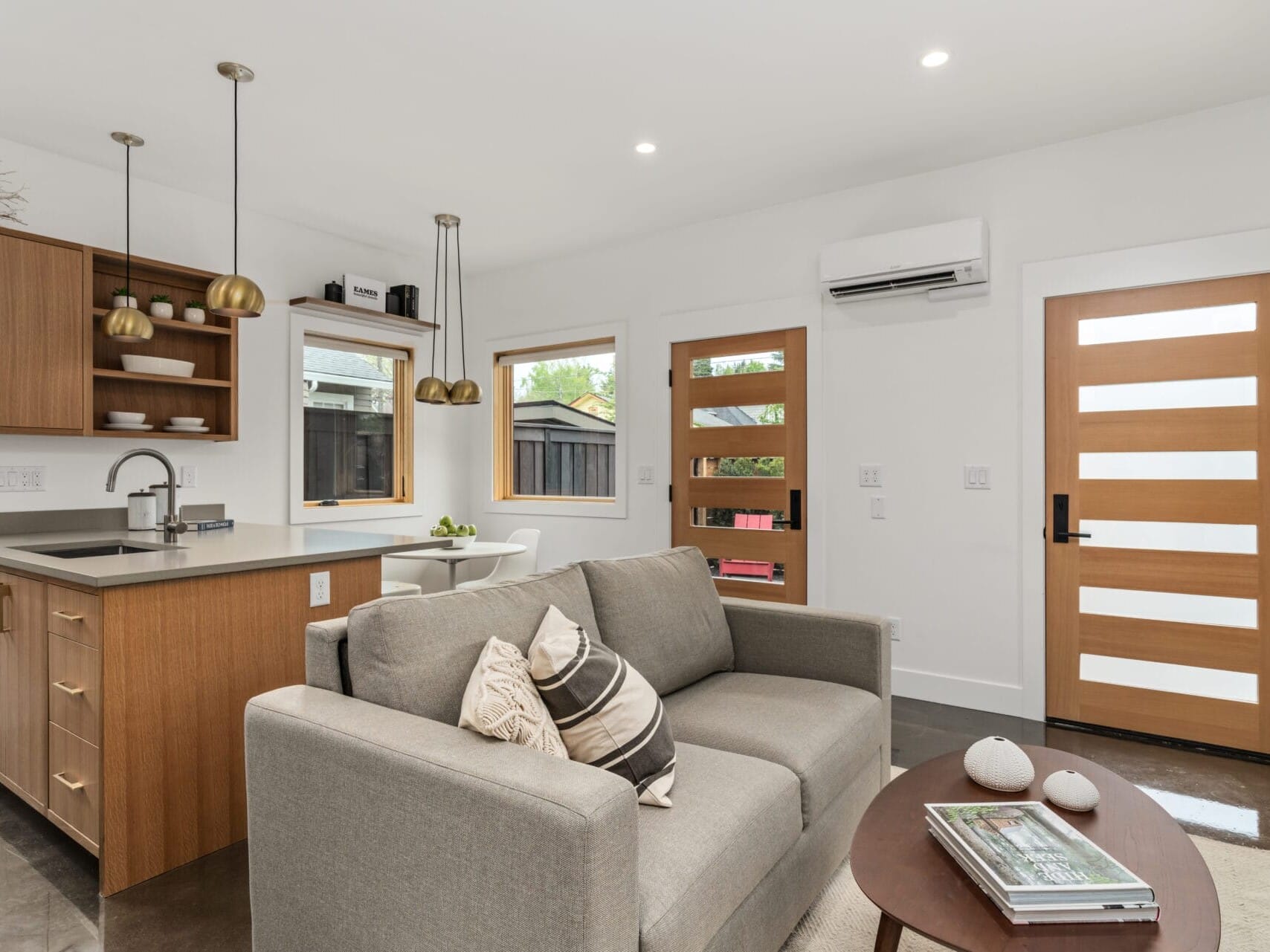 Modern living room and kitchen area in Portland, Oregon, with a gray couch, wooden coffee table, and pendant lights over a kitchen island. Natural light streams through two windows, complementing the wooden door with frosted glass panels. Decor includes books and small ornaments.