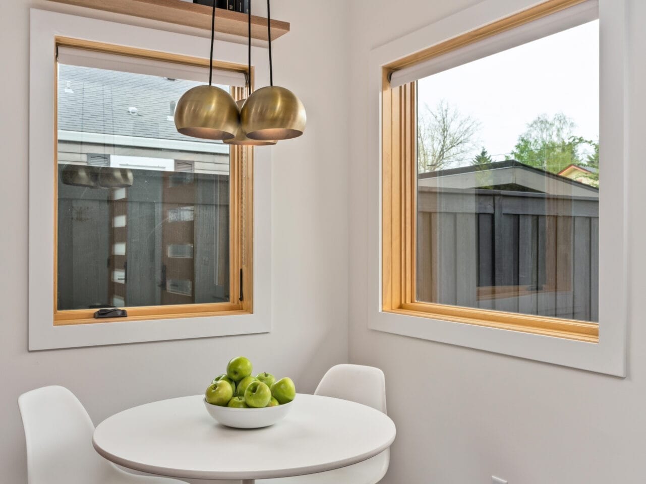 A small, modern dining nook in Portland, Oregon features a round white table with green apples in a bowl and two white chairs. Two large windows offer a garden view. A gold pendant light hangs above, while books and decor adorn the shelf over one window.