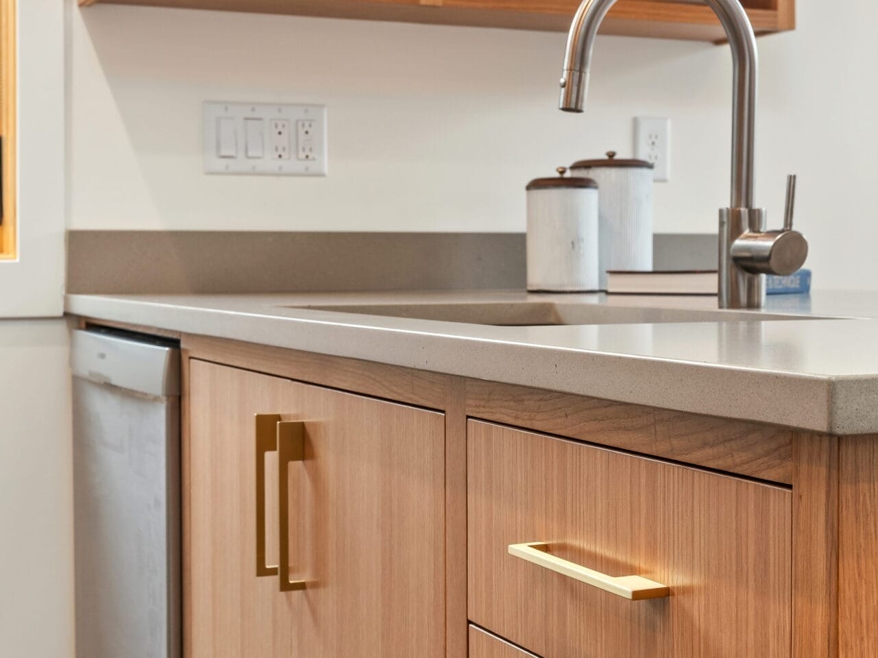 A modern kitchen in Portland, Oregon features a wooden cabinet with brass handles, a sleek countertop, and an open shelf holding white dishes. A stainless steel faucet is above the sink, with a dishwasher visible to the left.