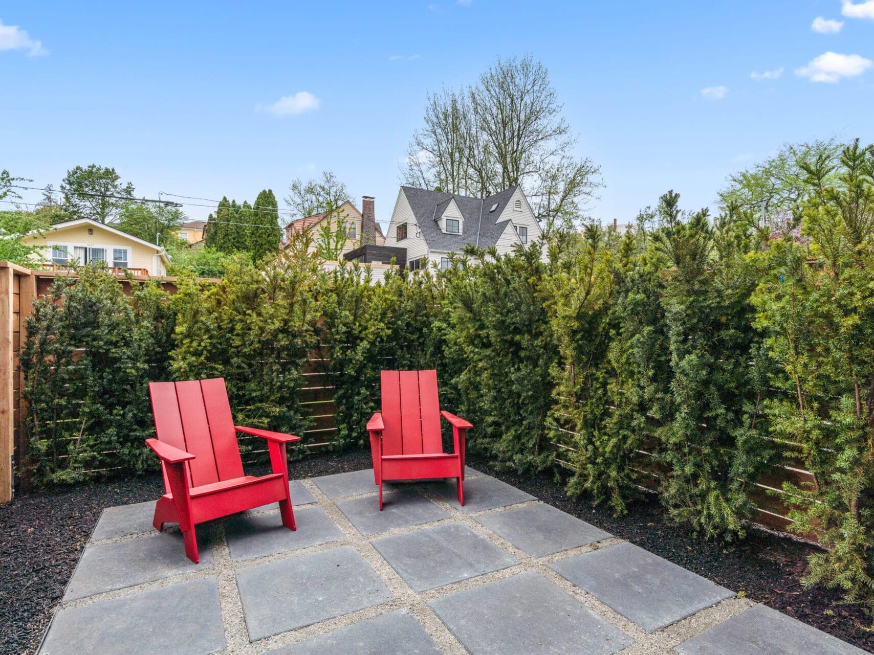 A backyard patio in Portland, Oregon features two red Adirondack chairs on a stone tile surface. The area is enclosed by tall evergreen shrubs, with residential houses visible against the blue sky.