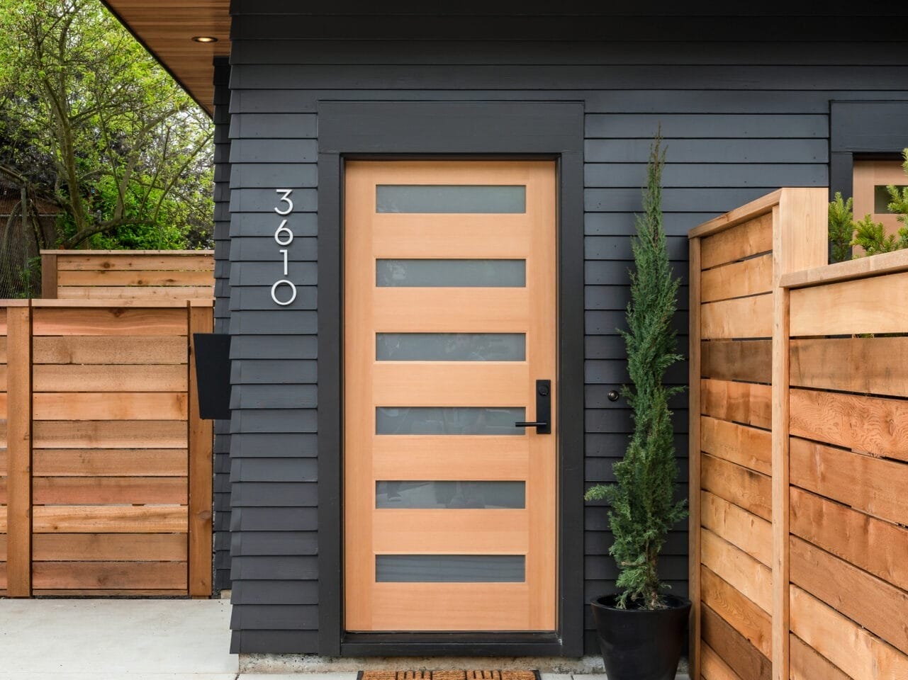 A modern house entrance in Portland, Oregon features a wooden door with horizontal glass panels. The exterior is dark gray with wooden accents. A small potted tree stands to the right, and the house number 3610 is displayed vertically beside the door.