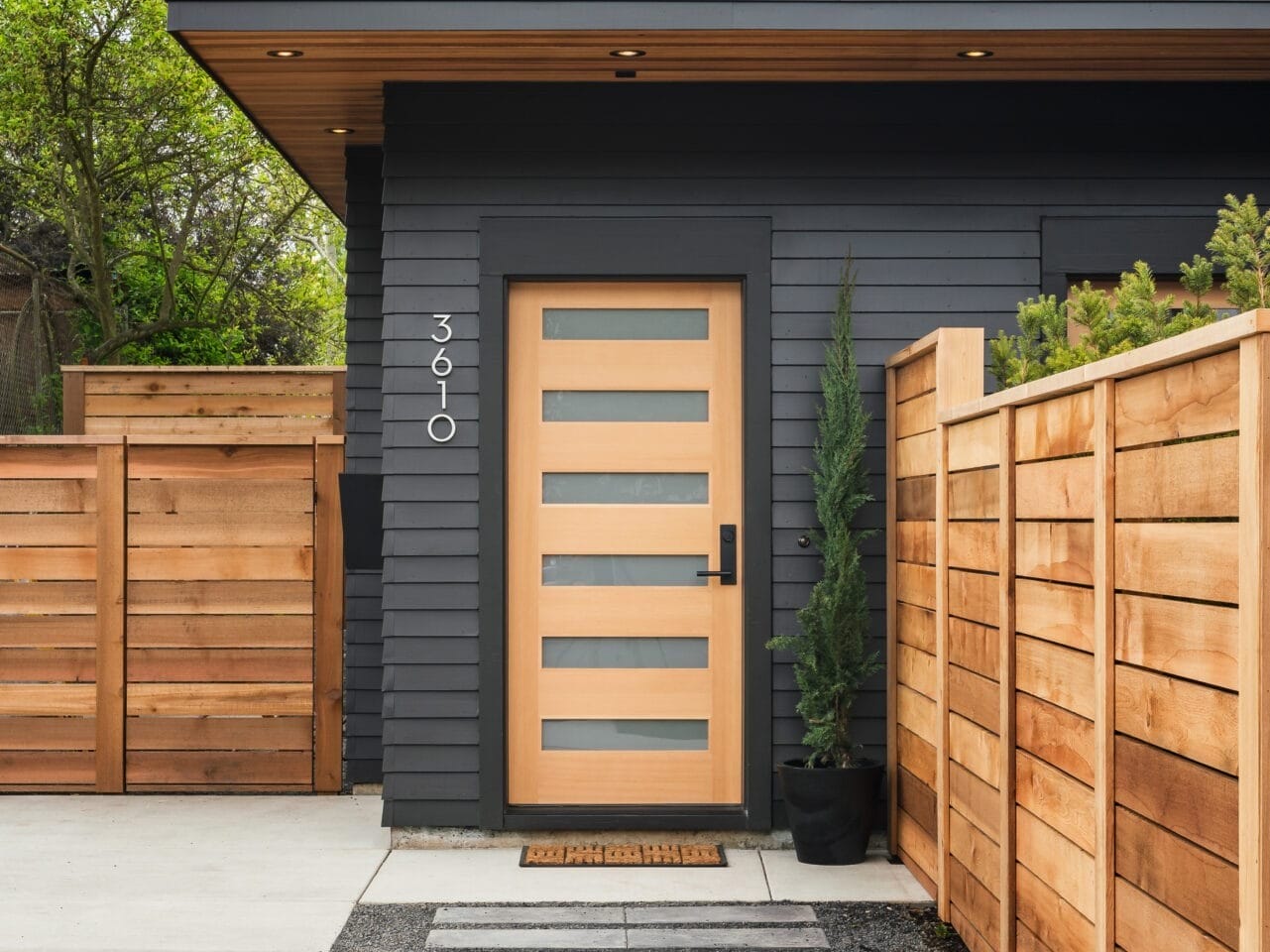 Modern house exterior in Portland, Oregon, with dark gray siding and a light wooden door featuring horizontal glass panels. A wooden fence lines the driveway, while a small tree adorns the entrance. Steps lead to the door from a paved walkway, with the house number clearly visible.