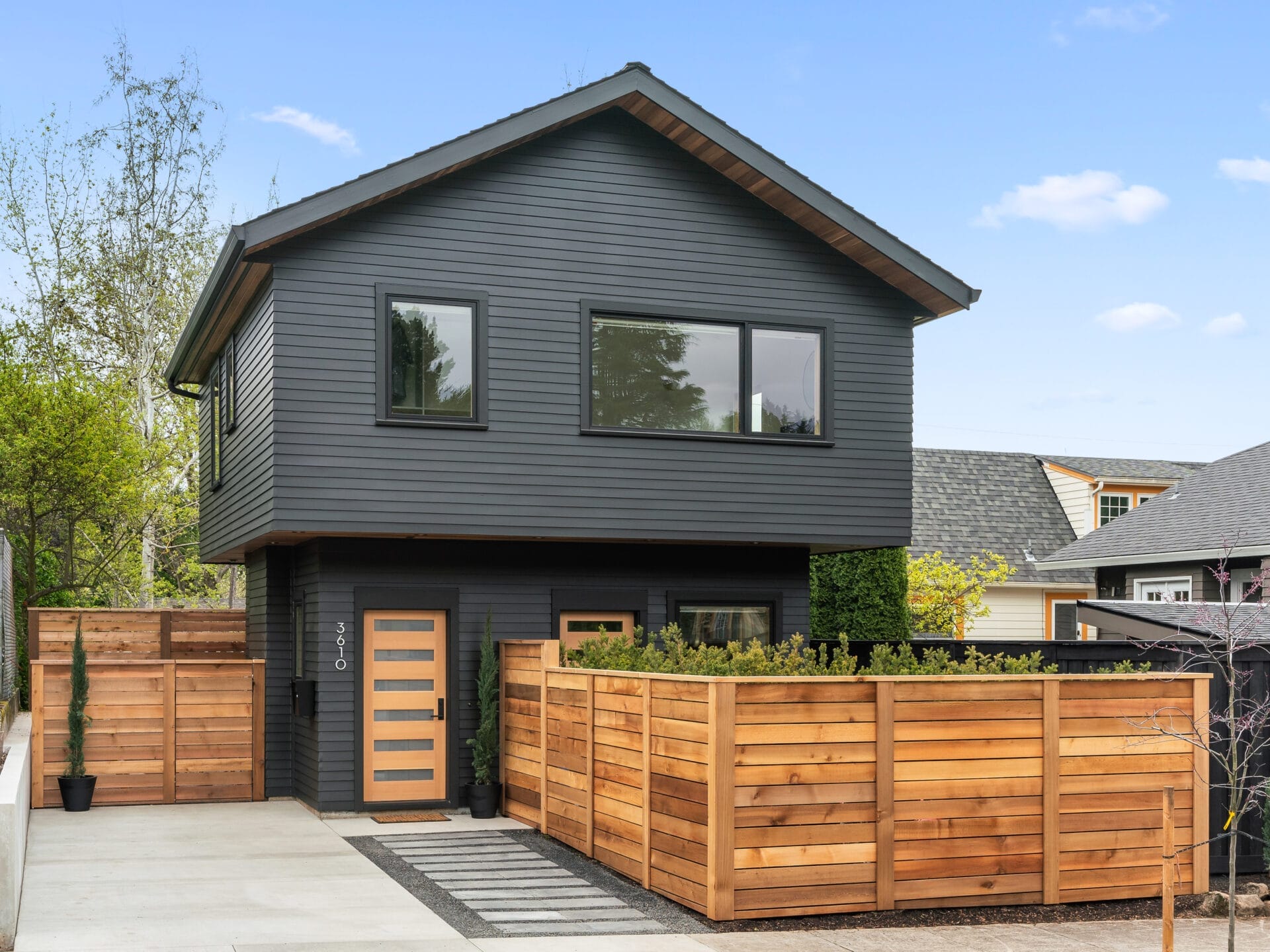 A modern two-story house in Portland, Oregon, features dark gray siding and large windows. A wooden fence surrounds the front yard, maintaining privacy. The minimalist design includes a few potted plants by the entrance, all under a clear blue sky.