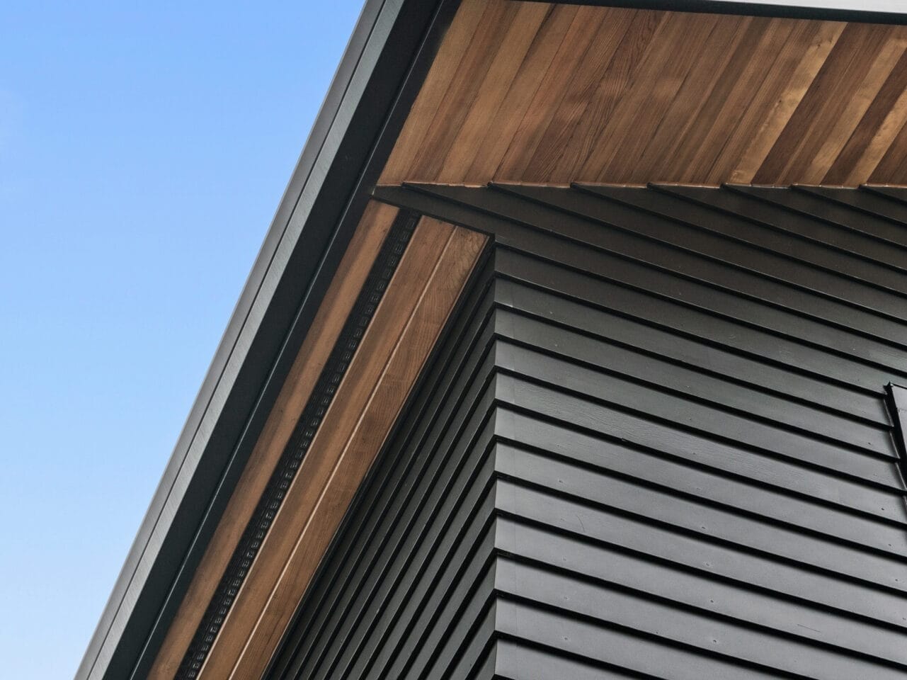 Close-up of a modern building's exterior in Portland, Oregon, featuring dark siding and wooden accents under the roof eaves. The structure is angled upwards against a clear blue sky.