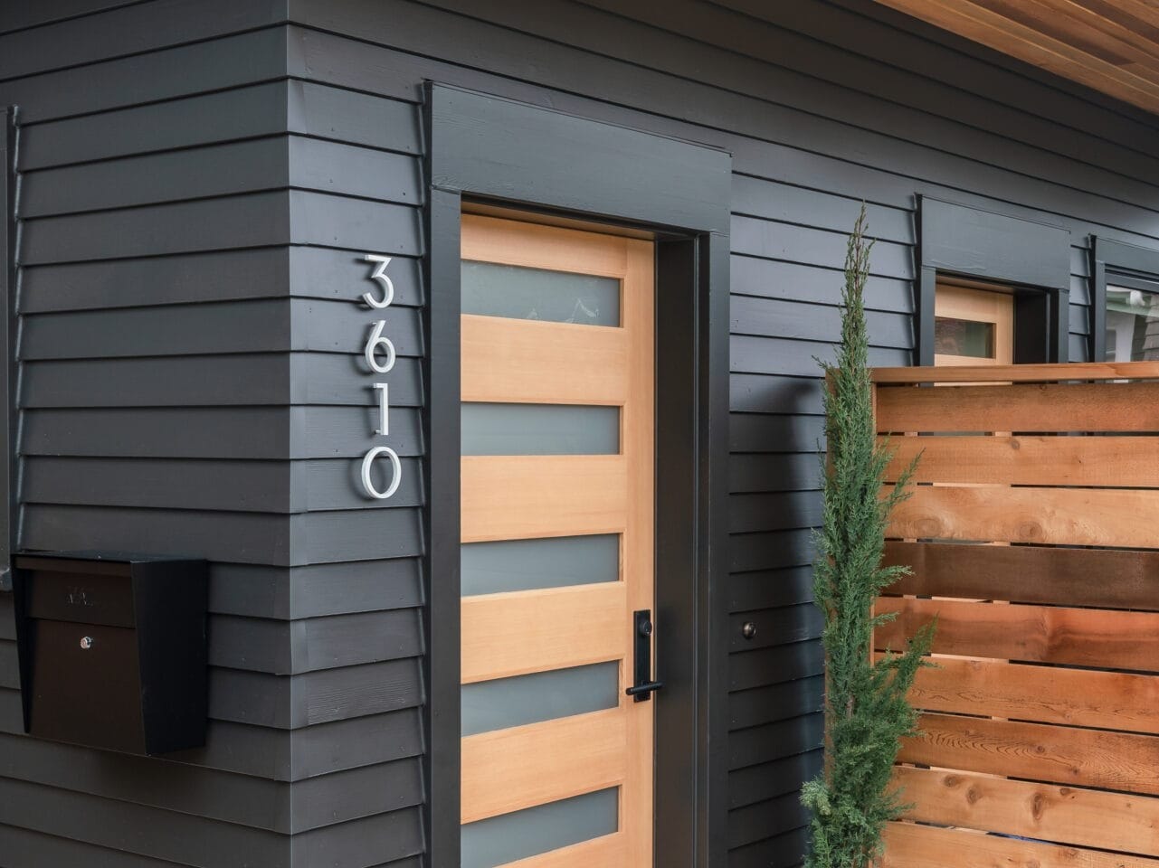 A modern house entrance in Portland, Oregon features a black and wood-themed door, numbered 3610. The exterior boasts horizontal black paneling and a wooden overhang. A potted plant is nestled near a wooden privacy screen beside the door, adding a touch of nature to this contemporary facade.