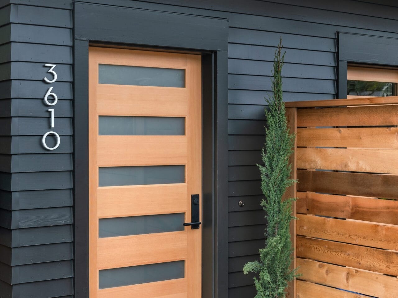 Wooden front door with horizontal glass panels on a modern dark-paneled house in Portland, Oregon. House number 3610 is displayed vertically. A potted plant sits beside the door, and a wooden privacy fence is partially visible. Two doormats adorn the concrete pathway.