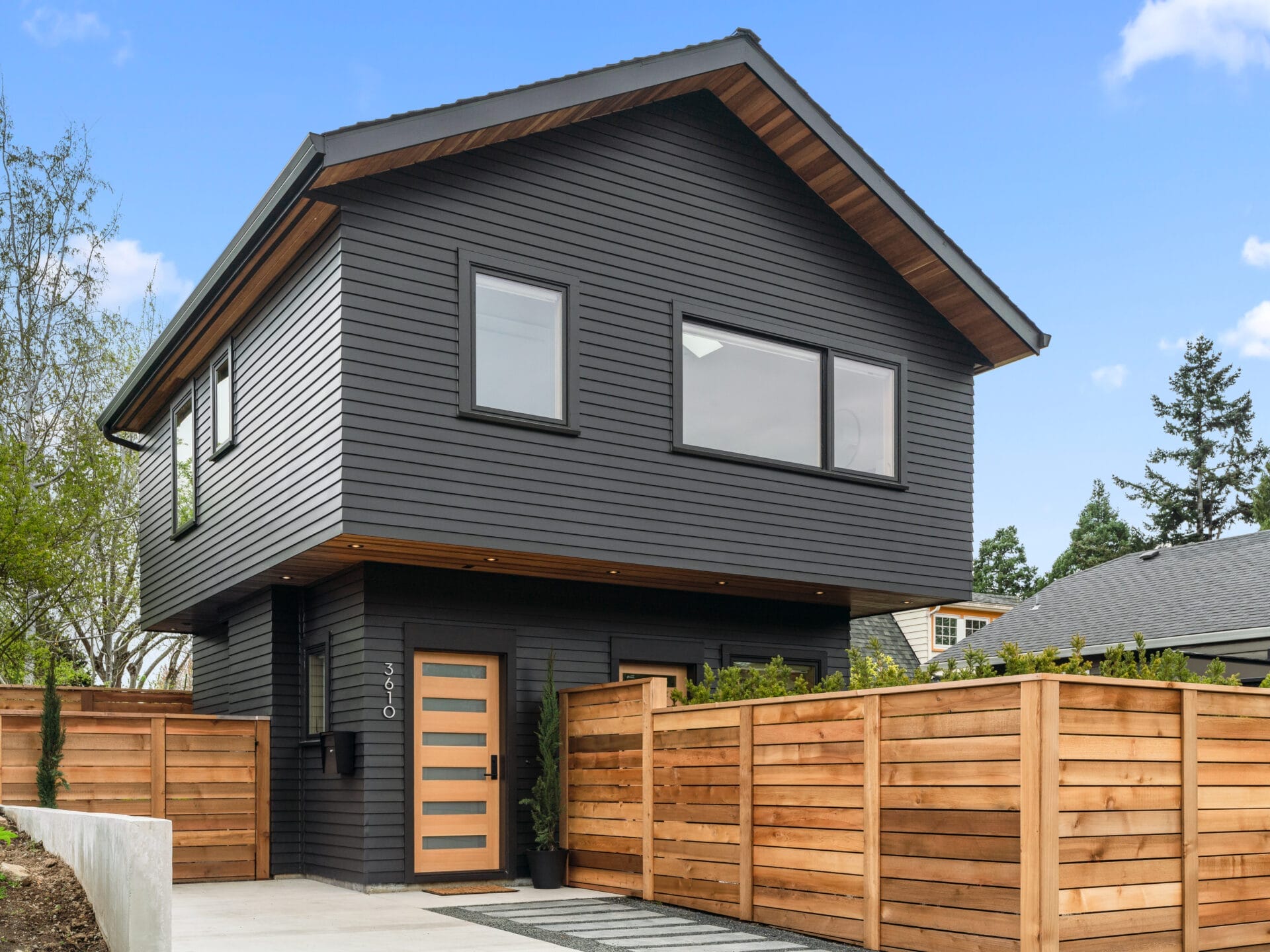 A modern two-story house in the heart of Portland, Oregon, boasts dark horizontal siding and a sloped roof. It features large windows and a sleek wooden door. A wooden fence encloses the property, with a paved driveway leading up to the entrance, framed by trees in the background.