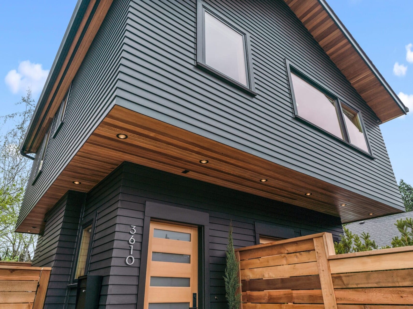 Modern two-story house in Portland, Oregon, featuring dark siding, large windows, and a wooden front door. The roof has a wood overhang, complemented by a wooden fence in the foreground. The address number 3610 is elegantly displayed next to the entrance.