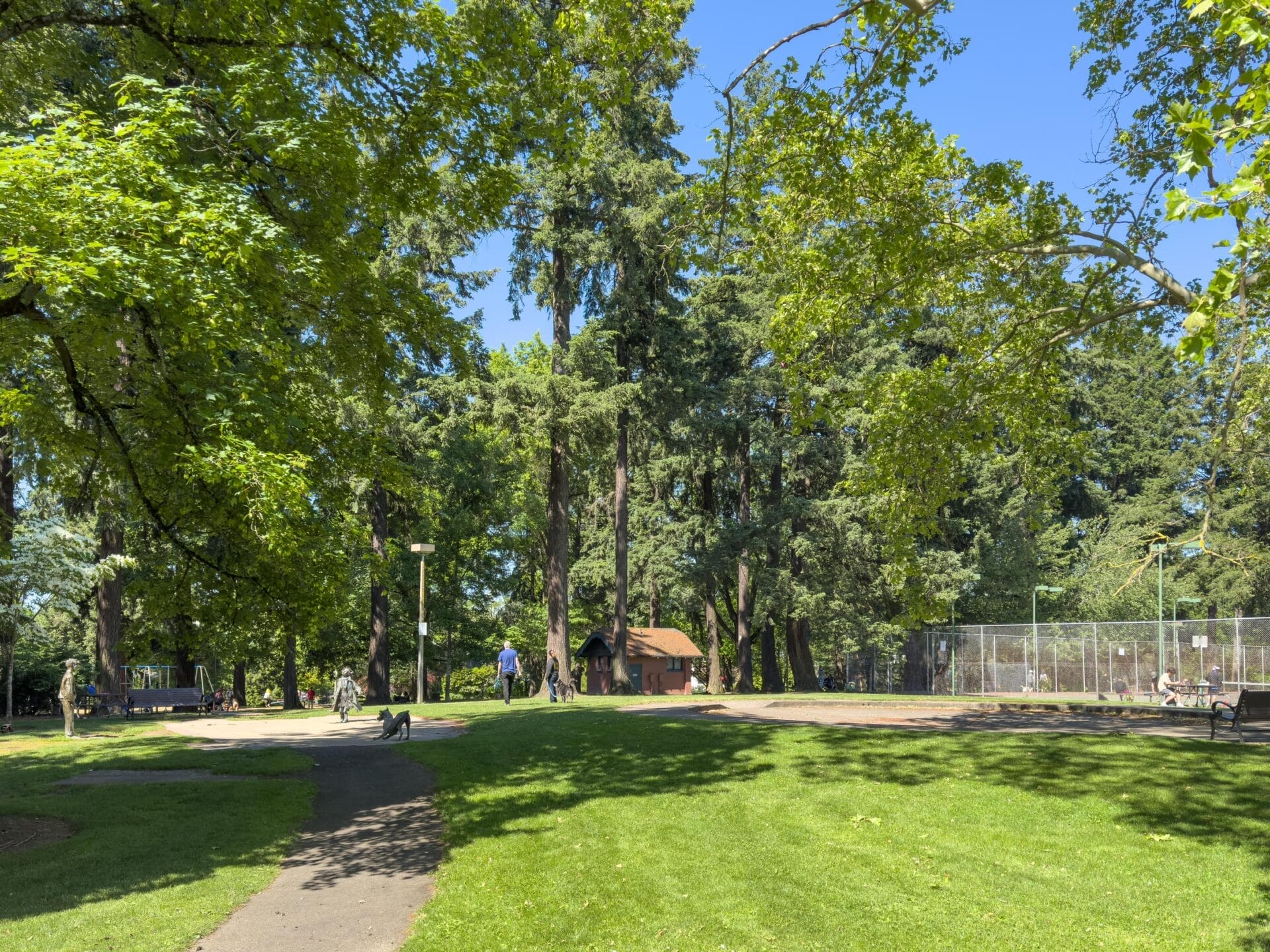 A sunny park scene with lush green grass and tall trees. A paved path winds through the park, where a few people walk and relax. A small building and a fenced tennis court are visible in the background.