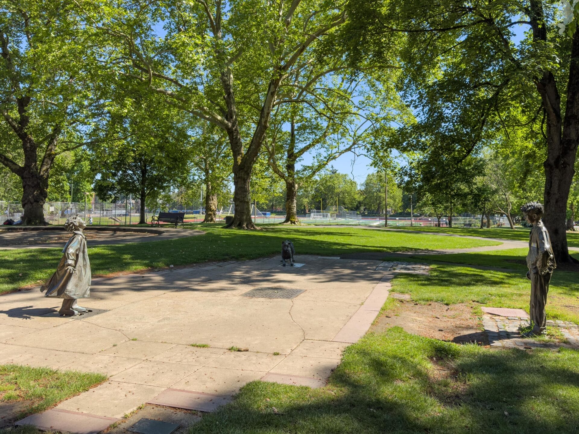 A park scene with two statues facing each other on a paved area surrounded by lush green grass and large trees. A dog statue is also present between them. In the background, there are tennis courts enclosed by fencing.