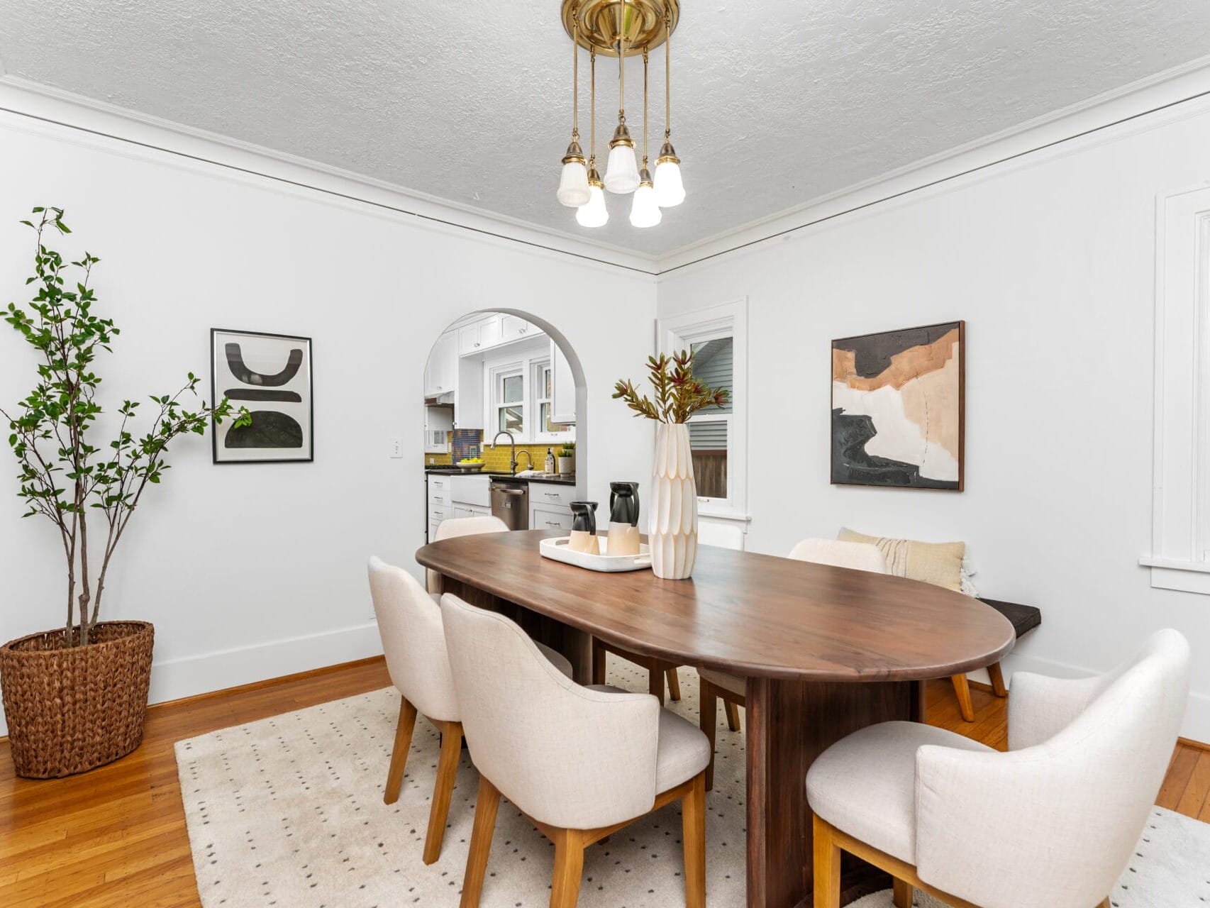 A modern dining room features a wooden oval table surrounded by six white upholstered chairs. Abstract art decorates the walls, and a basket with a tall plant sits nearby. An archway leads to a kitchen, and a pendant light hangs from the ceiling.