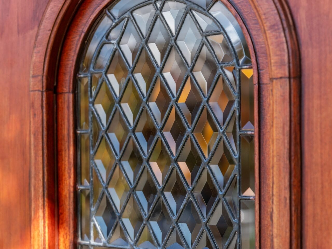 A wooden door features a small, arched stained glass window with a diamond pattern. The glass reflects light, creating a colorful effect. Warm sunlight casts soft shadows on the wood, enhancing the texture and details.