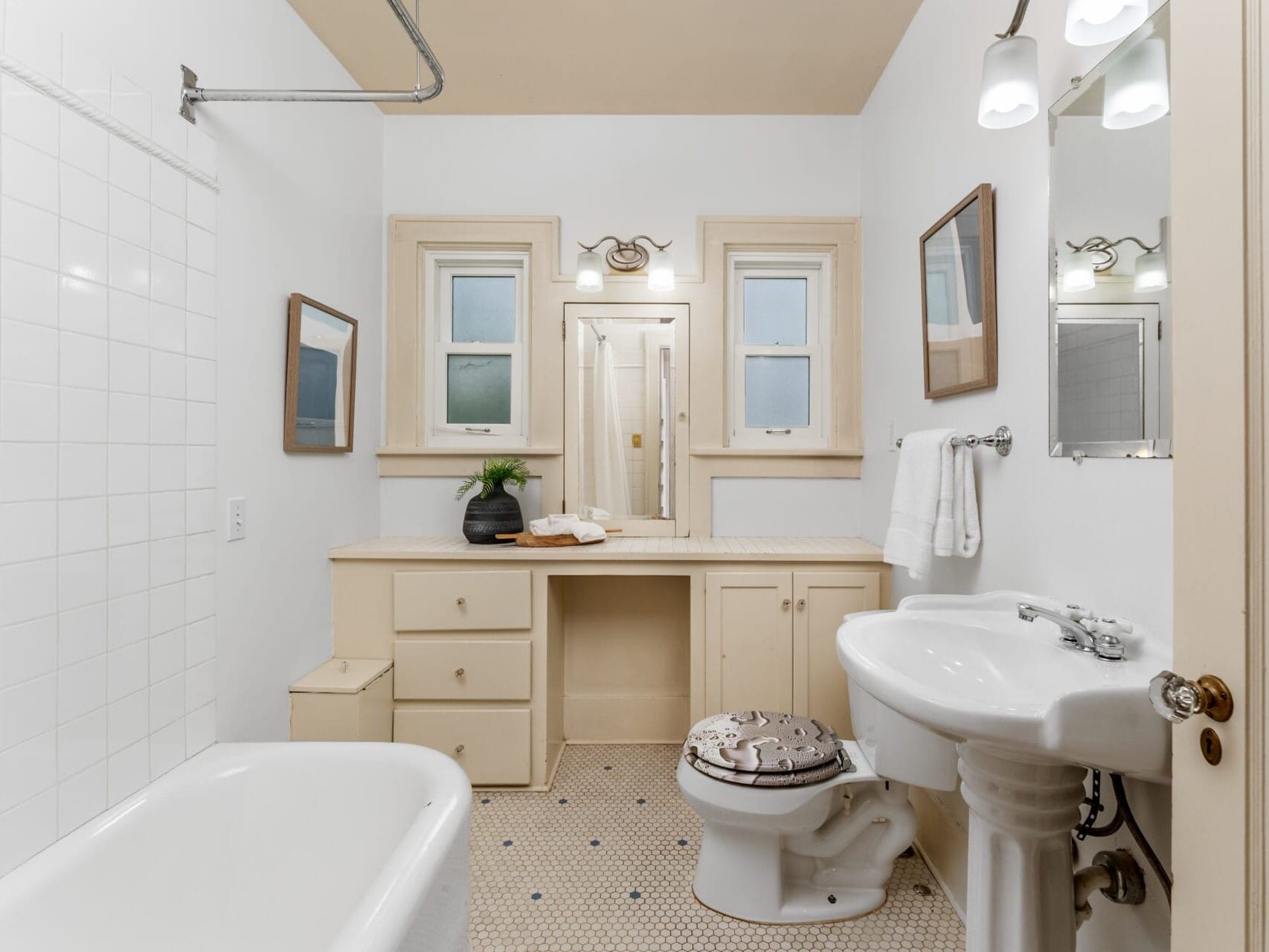 A bright bathroom with a white freestanding tub, pedestal sink, and toilet. There are two windows above a long beige vanity with drawers and a potted plant. The floor features small hexagonal tiles, and two wall lights illuminate the space.