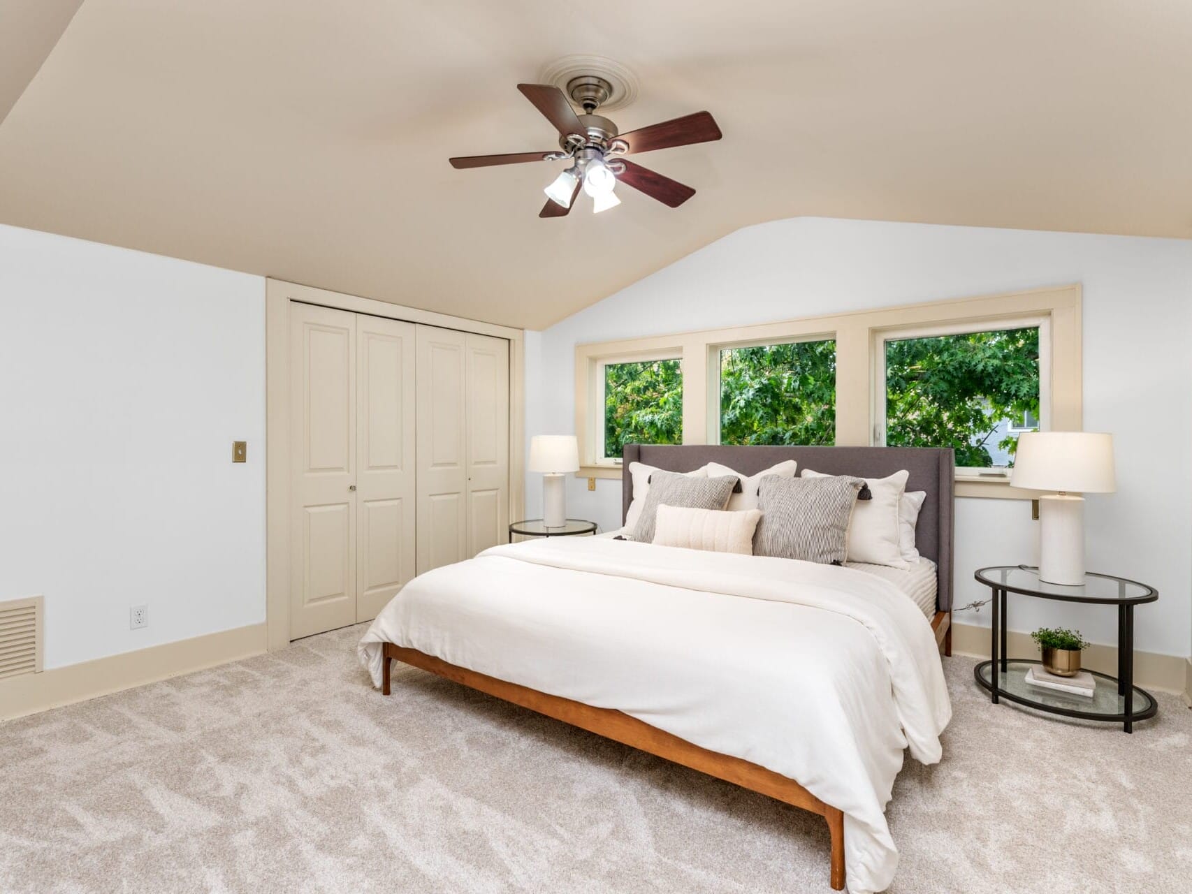 A cozy bedroom with a large bed featuring white and gray bedding. The room has a beige carpet, three windows with views of lush greenery, two bedside tables with lamps, a ceiling fan, and a double-door closet.