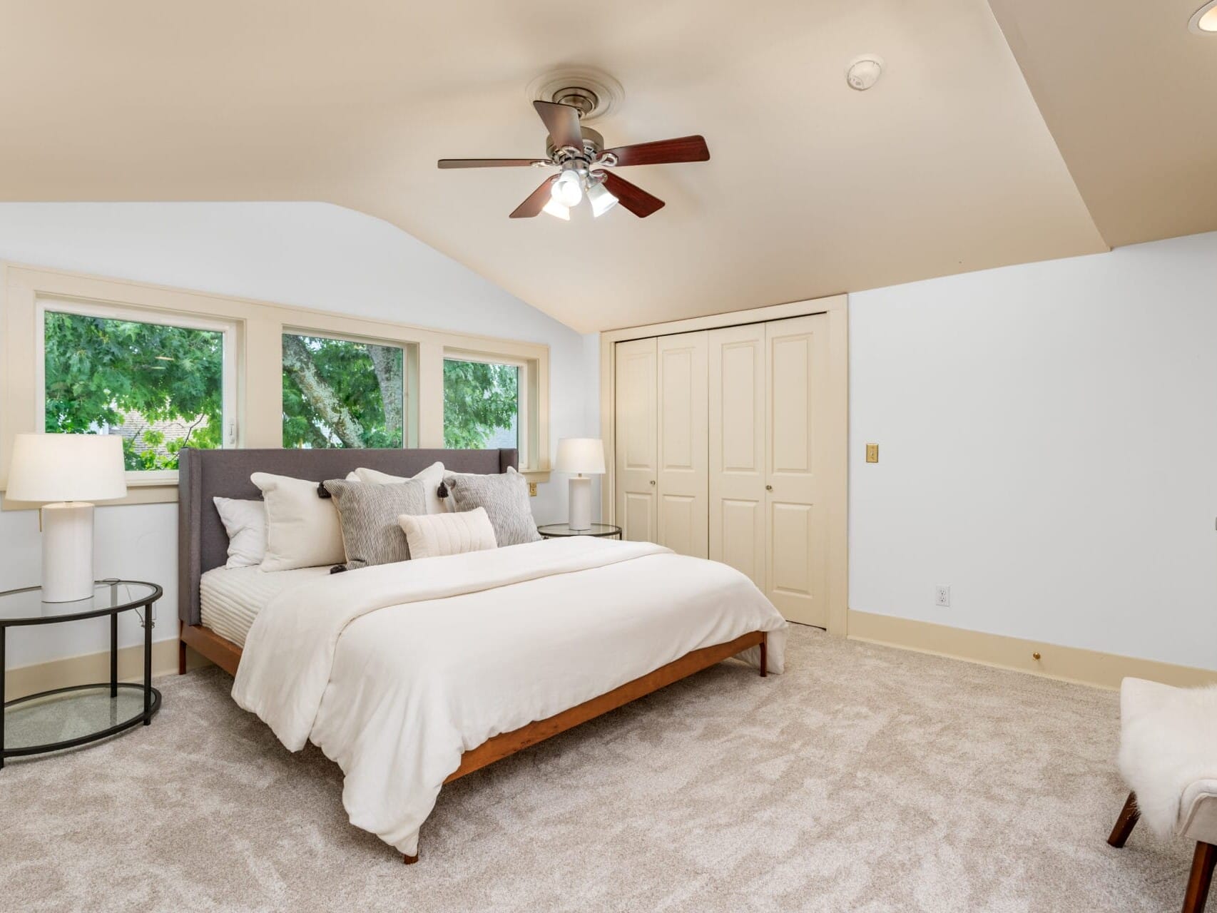 A bright, cozy bedroom with a large bed surrounded by white linens and plush pillows. Two round bedside tables with lamps flank the bed. Theres a ceiling fan above, and natural light streams in through three windows. A closet is in the background.