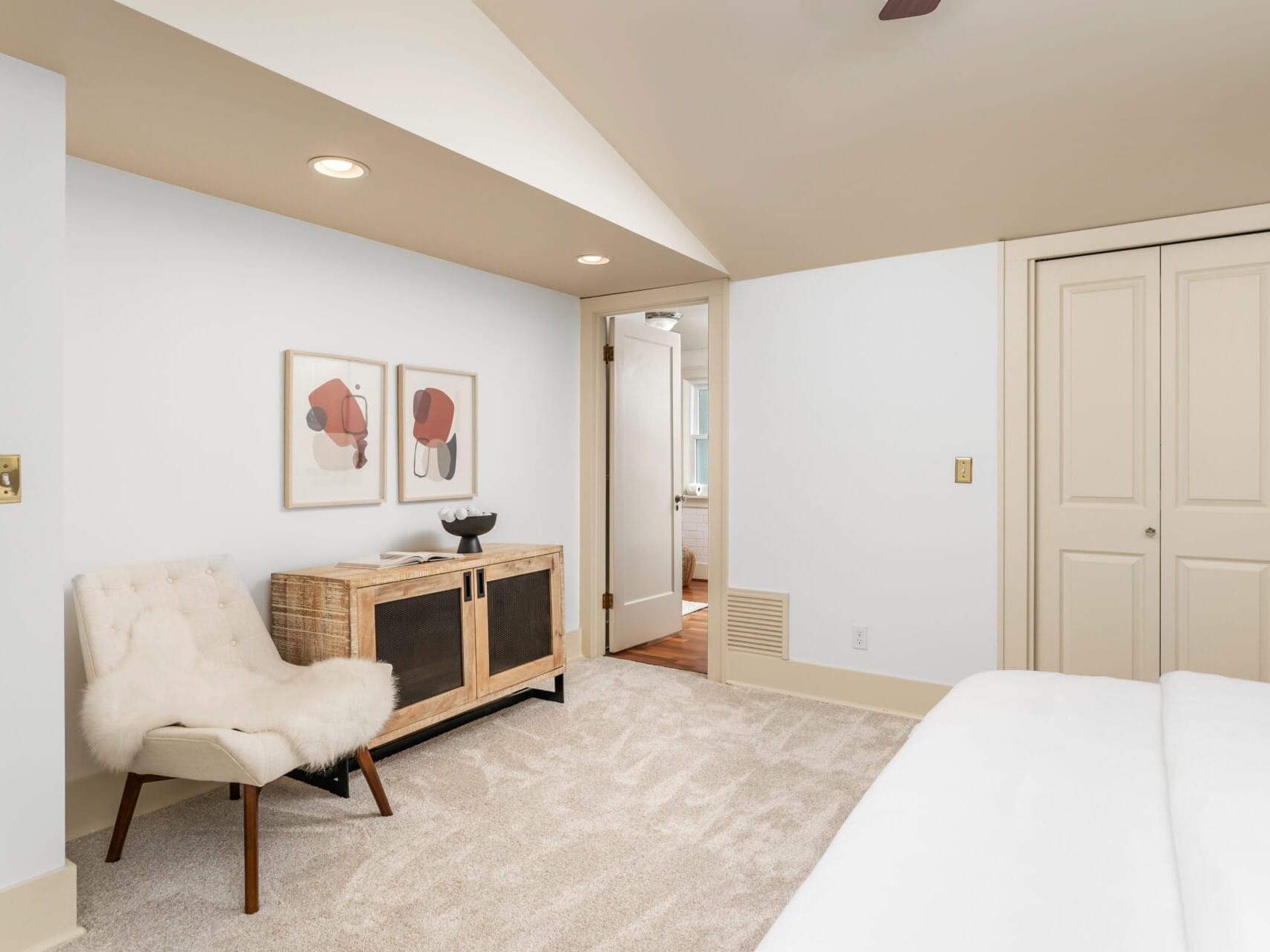 A minimalist bedroom with a cozy chair, a wooden cabinet, and framed abstract art on the wall. Beige carpet covers the floor, and theres a closet with sliding doors. Soft lighting creates a warm, inviting atmosphere.