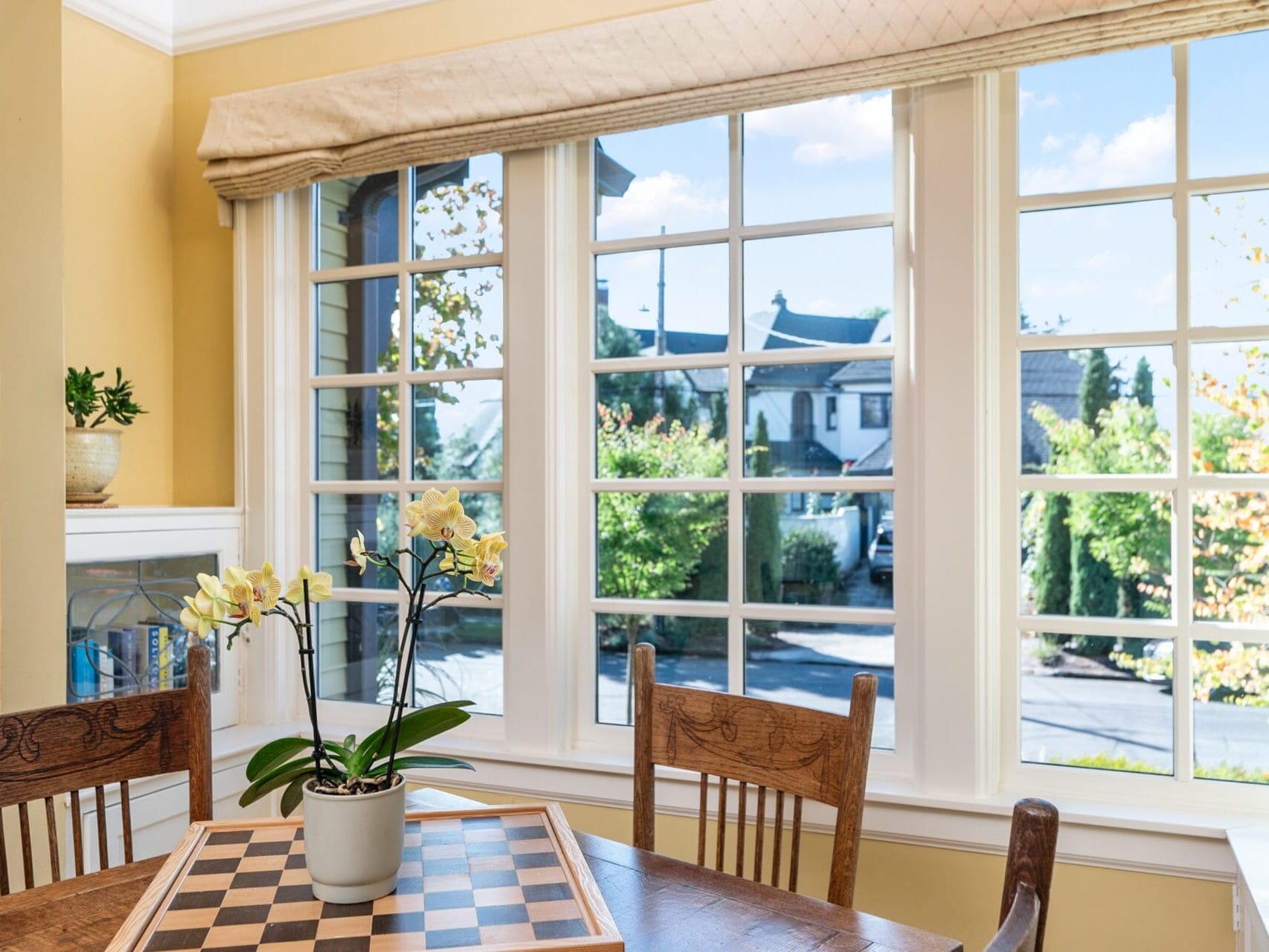 A dining area with wooden chairs and a table featuring a chessboard and potted orchids. Large windows provide a view of houses and a clear blue sky outside. Light yellow walls and a curtain add warmth to the room.