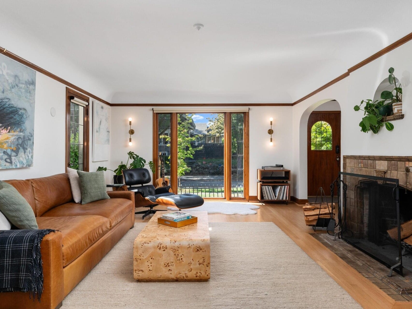 A cozy living room with a brown leather sofa, cushioned with white and gray pillows, faces a fireplace. Large windows and a wooden door open to a green garden. Modern art and a black armchair with an ottoman accent the space.