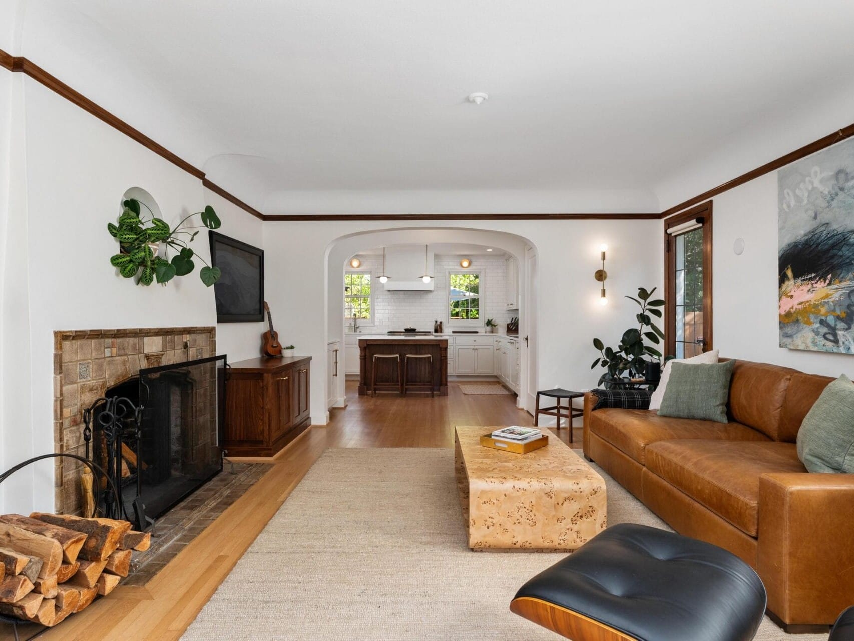 A cozy living room with a brown leather sofa, a wooden coffee table, and a fireplace with stacked logs. Theres a large painting on the wall, plants, and a view into a white kitchen with dark cabinets. Natural light fills the space.