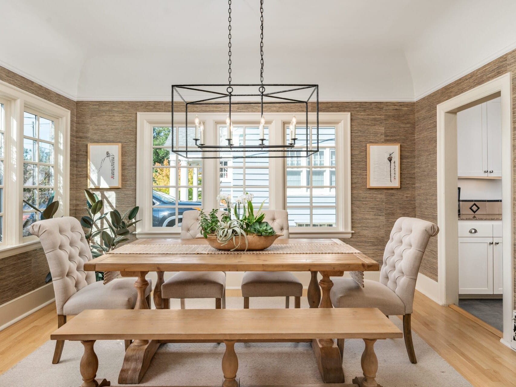 A dining room featuring a wooden table with a centerpiece of green plants surrounded by four upholstered chairs and a wooden bench. A modern chandelier hangs above, and light streams in through multiple windows. Neutral tones decorate the space.