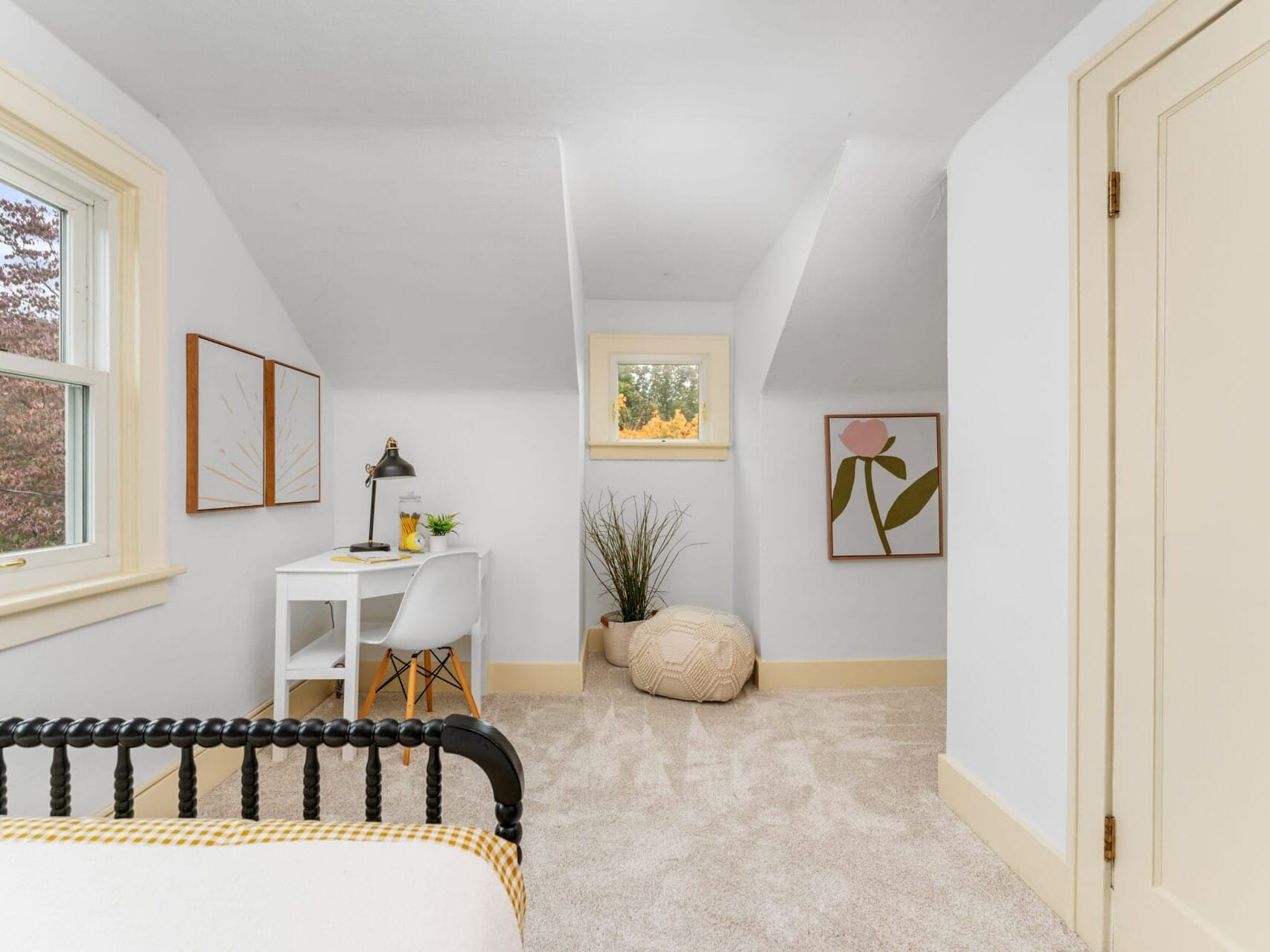 A cozy attic bedroom with sloped ceilings featuring a small desk by a window, a black spindle bed, and minimalist artwork on the walls. A pouf and plant sit near a second window. Soft, neutral tones enhance the tranquil atmosphere.