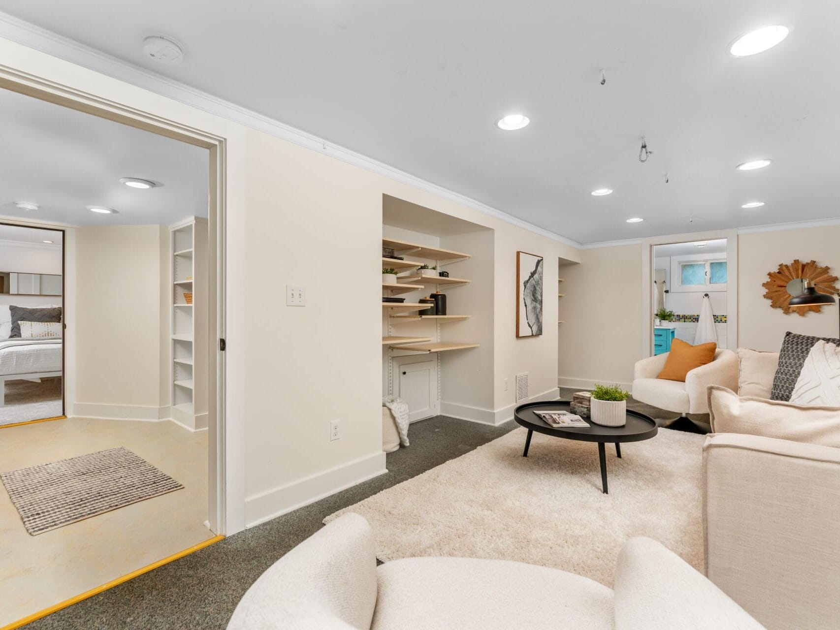 A cozy living room with white walls and carpet, featuring a beige sofa, round black coffee table, and decorative items. An open doorway leads to a bedroom with a neatly made bed. Recessed ceiling lights illuminate both areas.