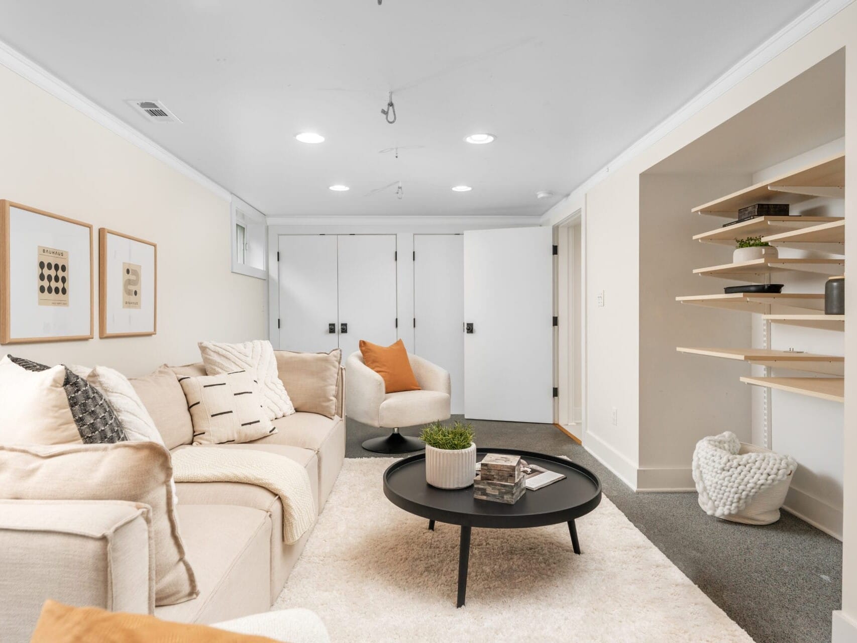 A cozy living room featuring a beige sectional sofa with assorted pillows, a round black coffee table with a plant, and built-in shelves. Neutral-toned wall art decorates the light-colored walls, and recessed lighting brightens the space.