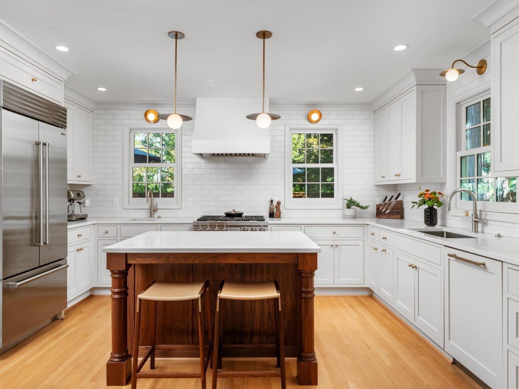 A modern, bright kitchen with white cabinets, wood flooring, and a central island with two stools. Stainless steel appliances and three pendant lights hang above the island. Two windows provide natural light, and a vase with flowers sits on the counter.