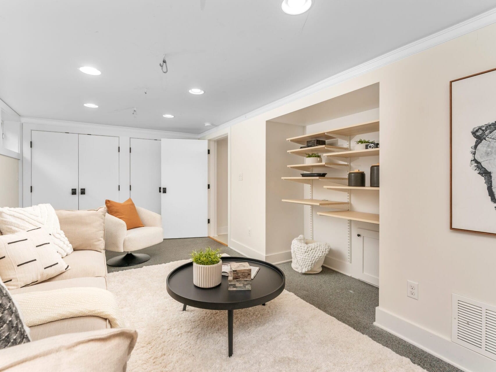 Cozy living room with a beige sectional sofa and a round black coffee table on a white rug. A modern art piece hangs on the wall to the right. Built-in shelves hold decorative items, and double white doors are visible in the background.