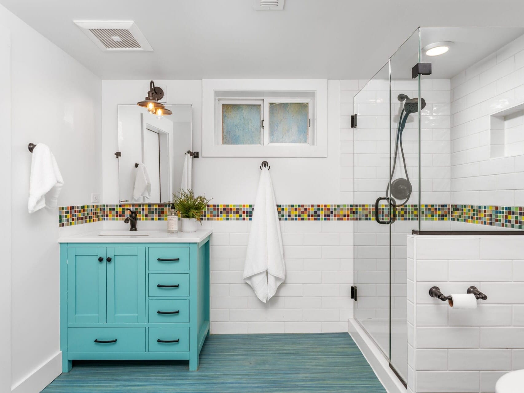 Modern bathroom with a turquoise vanity featuring a white countertop and a round mirror. The walls are white with a colorful mosaic tile border. A glass-enclosed shower is on the right, and theres a towel rack with a white towel.