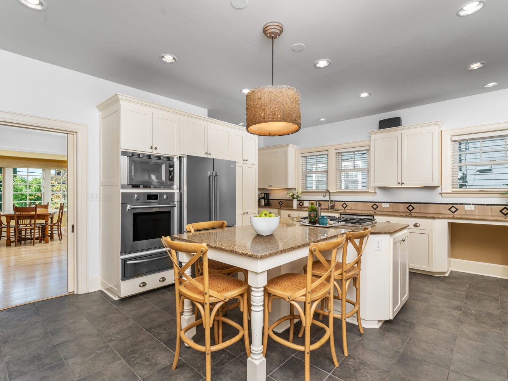 A spacious, modern kitchen with white cabinets, a large island with a granite countertop, and four wooden barstools. Stainless steel appliances and a pendant light hang above the island. A dining area is visible through an open doorway.