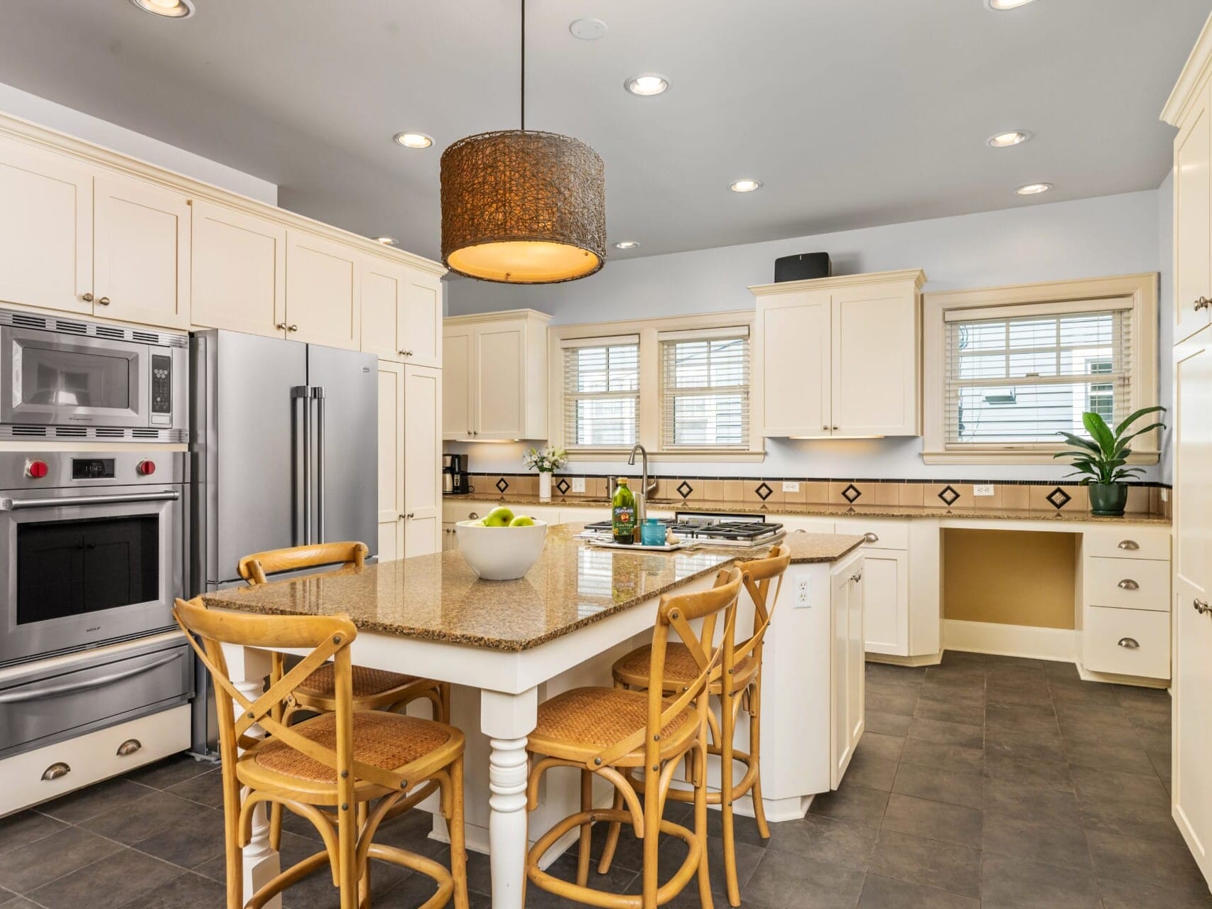 A modern kitchen with white cabinets, a stainless steel double oven, and a large refrigerator. An island with a granite countertop and four wicker chairs is centered. A fruit bowl and a bottle are on the island. Large windows provide natural light.