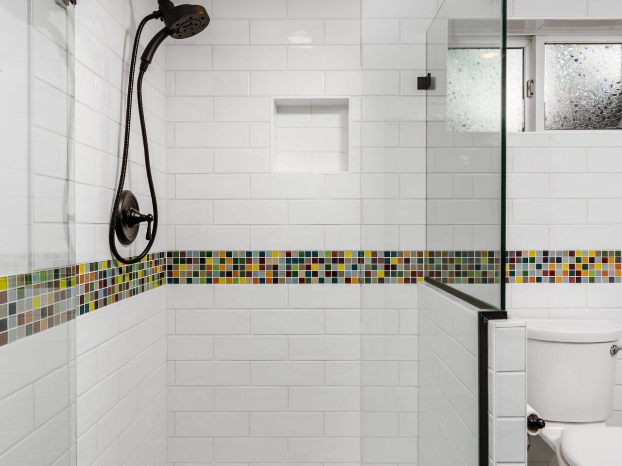 Modern bathroom with a glass-enclosed shower featuring a black showerhead. Walls are lined with white subway tiles and a horizontal strip of colorful mosaic tiles. There is a small window above a white toilet, and the floor has blue tiles.