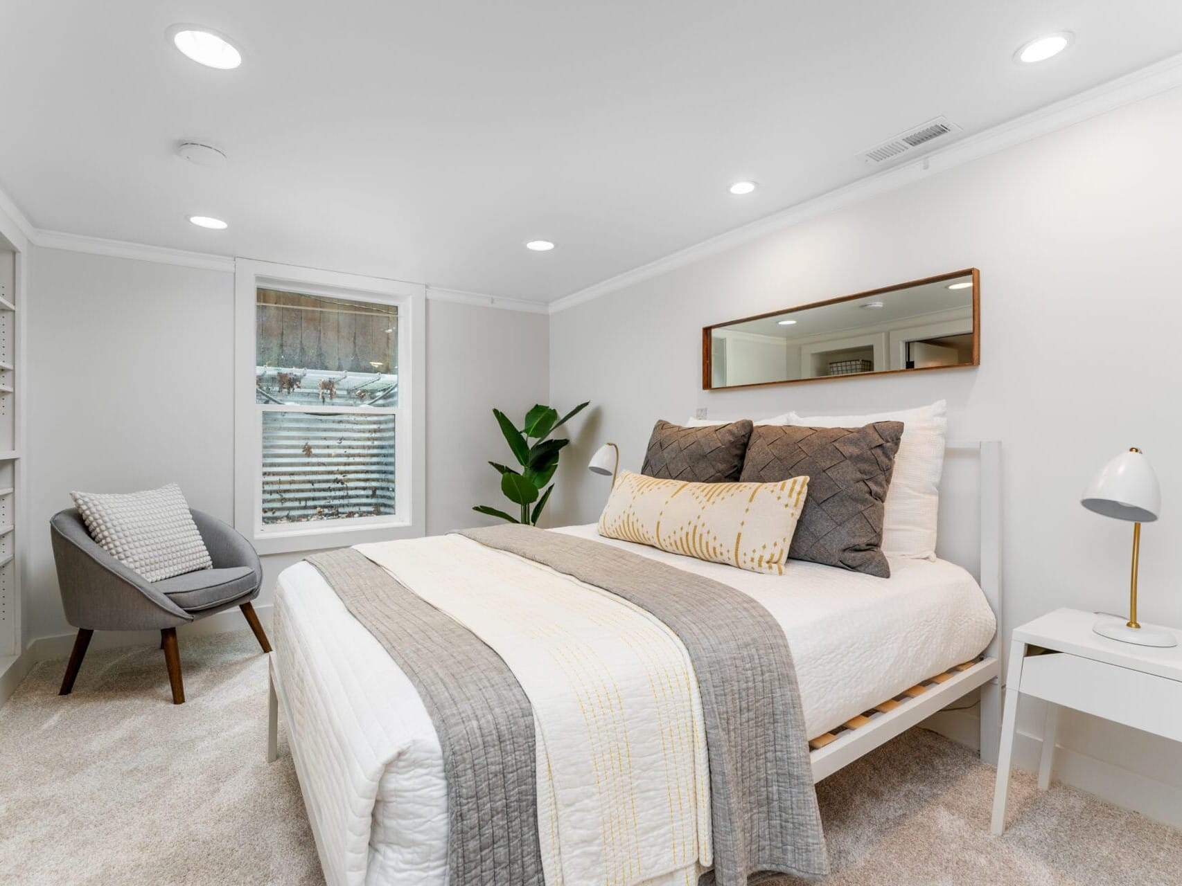 A bright bedroom with a neatly made bed featuring gray and white bedding. Theres a modern chair in the corner, a potted plant near the window, and a small white nightstand with a lamp. A rectangular mirror hangs above the bed.