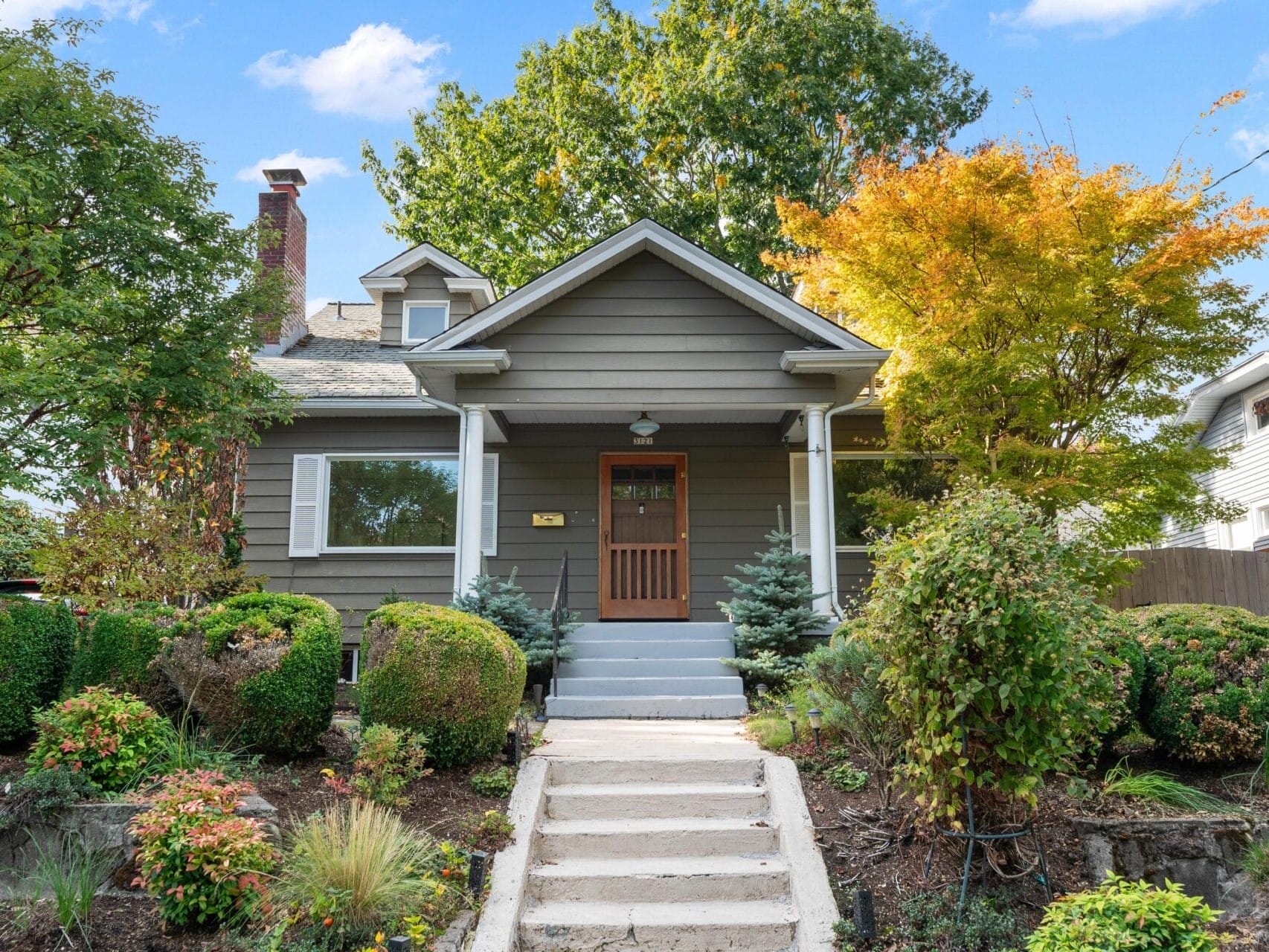 A charming, gray single-story house with a small porch and a wooden door. Its surrounded by colorful trees and neatly trimmed bushes. A concrete pathway leads up to the entrance, and the sky is clear with a few clouds.