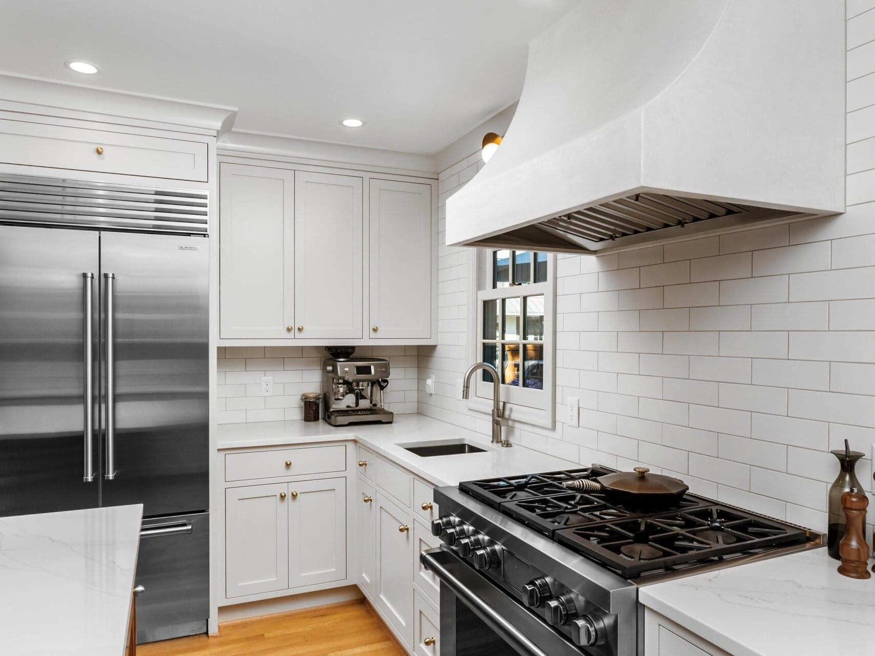 Modern kitchen with white cabinets, a stainless steel refrigerator, and a gas stove with a large white range hood. The countertop has a coffee machine and various decor items. White subway tiles line the walls, and wood flooring completes the look.