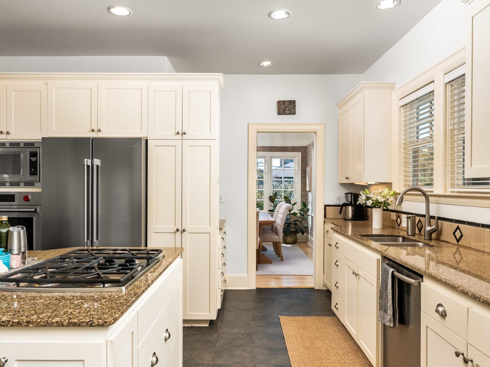 A modern kitchen with cream-colored cabinets, a granite countertop island with a gas stovetop, and stainless steel appliances. A doorway leads to a dining area in the background, with bright lighting from ceiling fixtures.