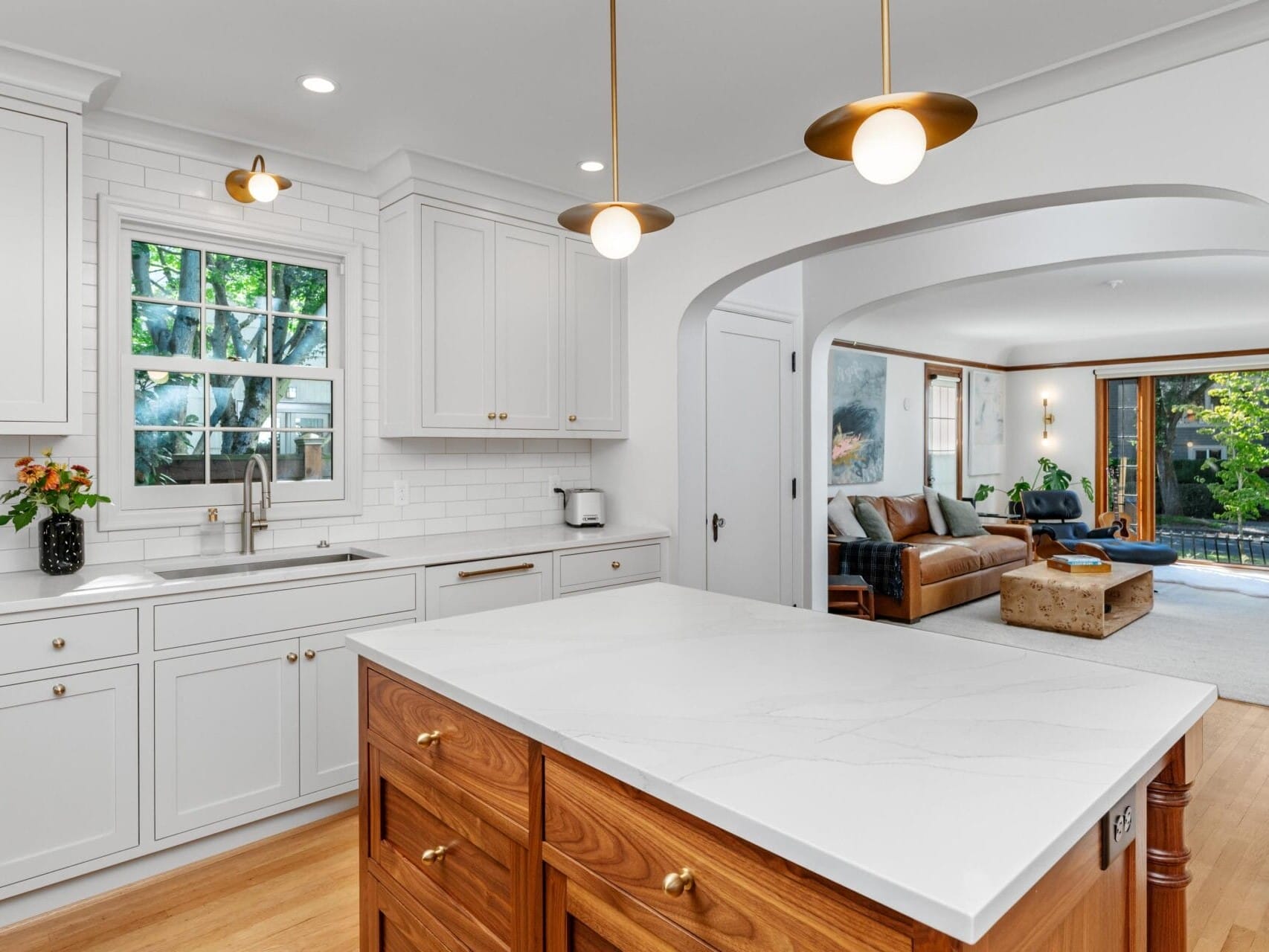 A modern kitchen with white cabinets, marble countertop, and wooden island. Gold pendant lights hang above. The kitchen opens to a cozy living area with a blue sofa and large windows overlooking a garden.