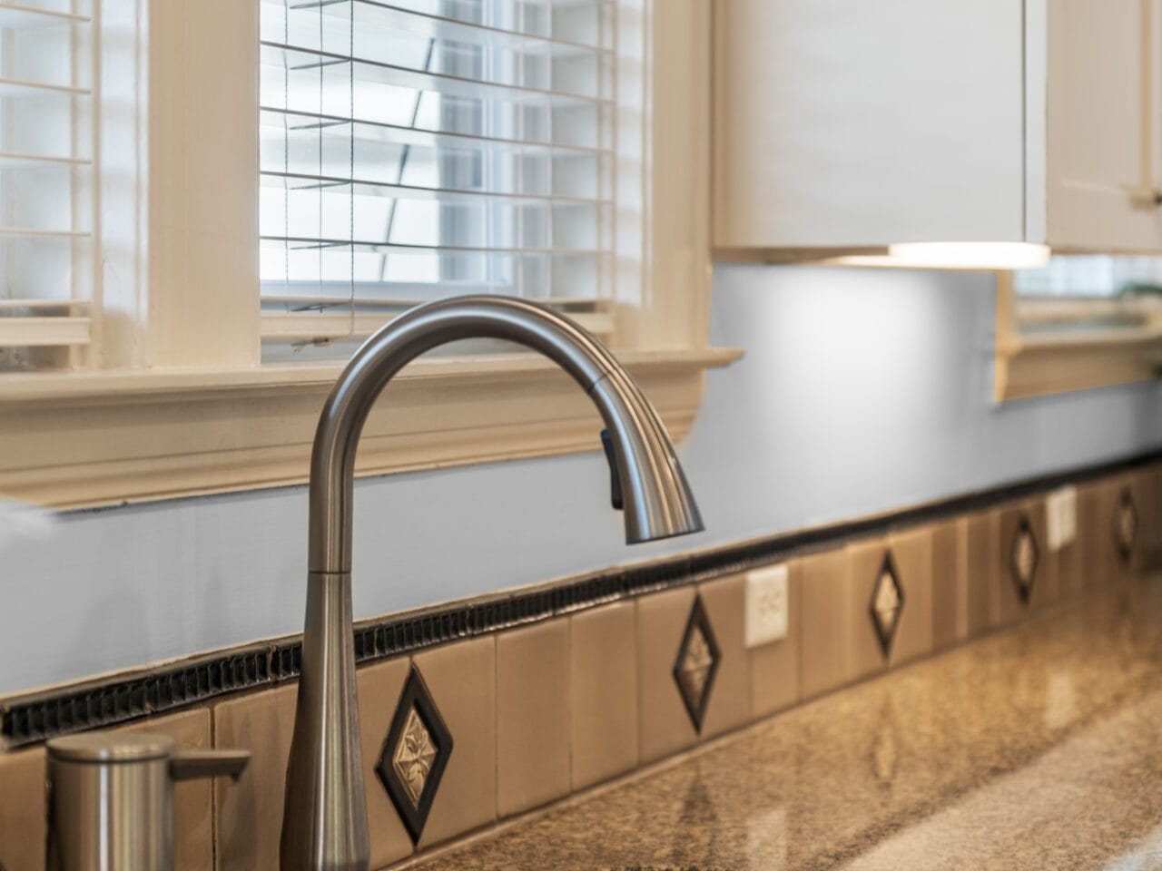 A modern kitchen sink with a sleek, curved faucet is set into a polished granite countertop. The backsplash features square and diamond-shaped tile patterns, and a window with white blinds allows natural light to illuminate the space.