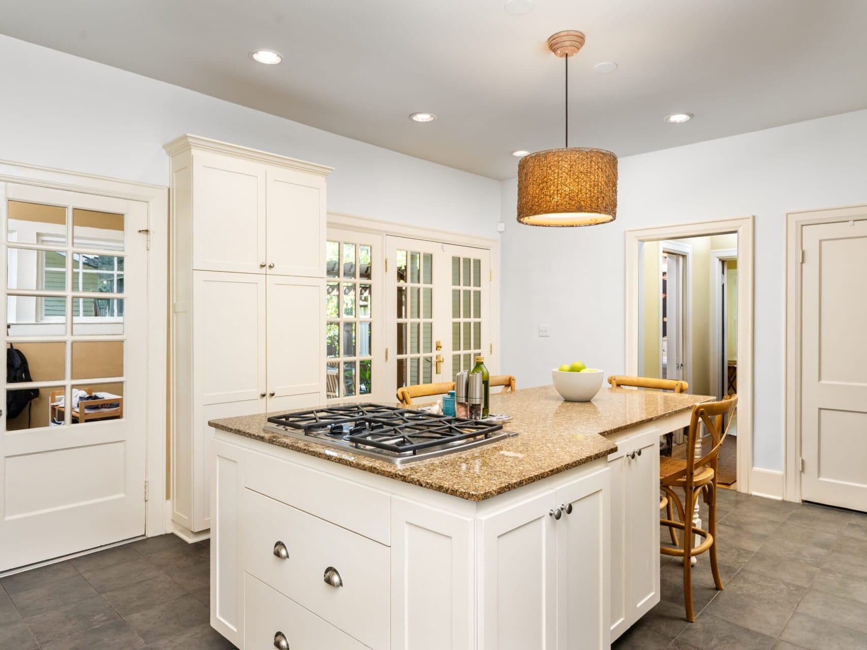 A modern kitchen with a large island featuring a built-in stove and granite countertop. Wooden chairs surround the island, and a fruit bowl sits on top. The room has white cabinets, a pendant light, and multiple doors and windows.