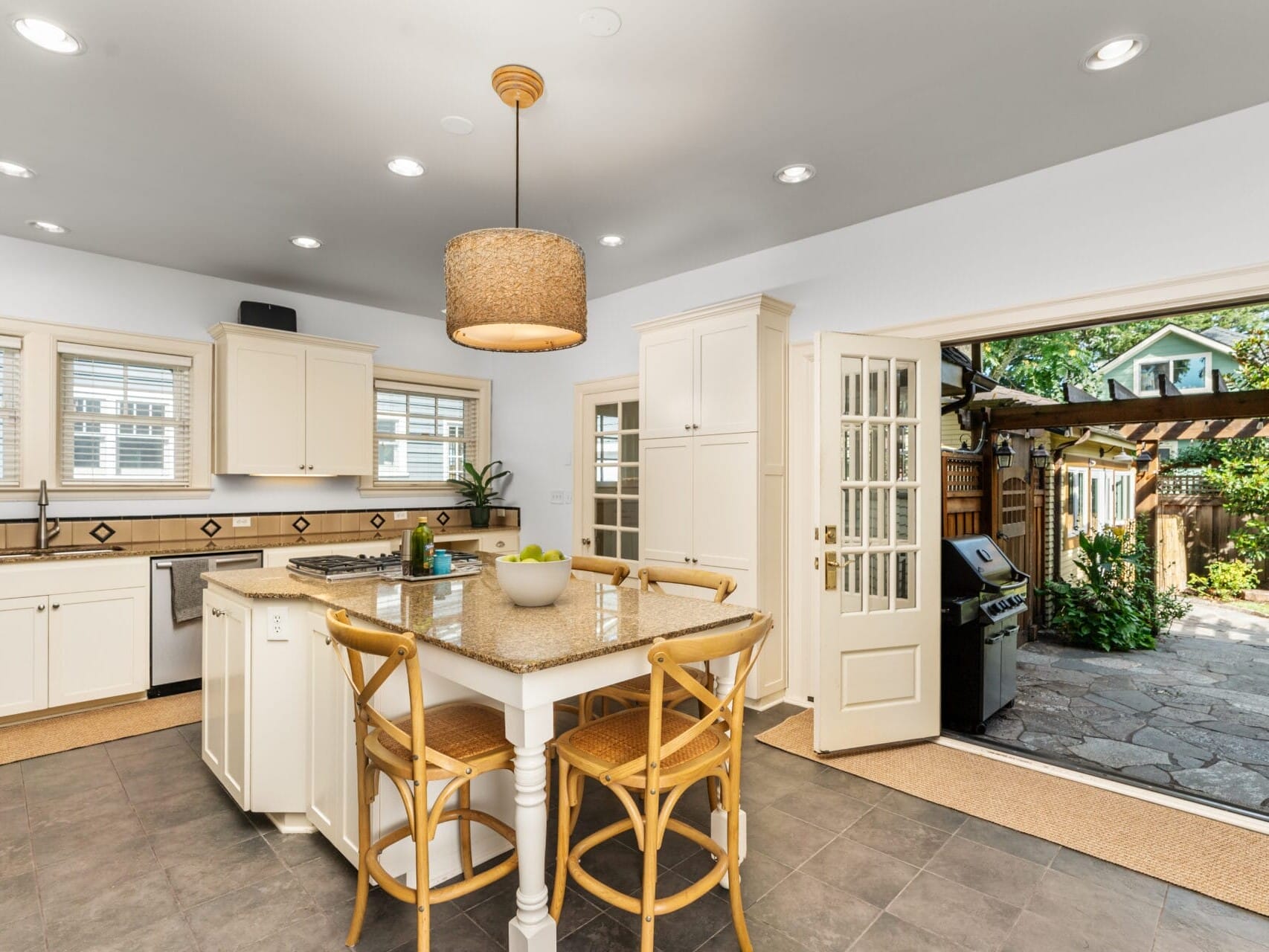 A bright kitchen with white cabinets and a central island featuring four wooden chairs. A large windowed door opens to a patio with a grill and pergola, revealing a garden with greenery. A woven pendant light hangs above the island.